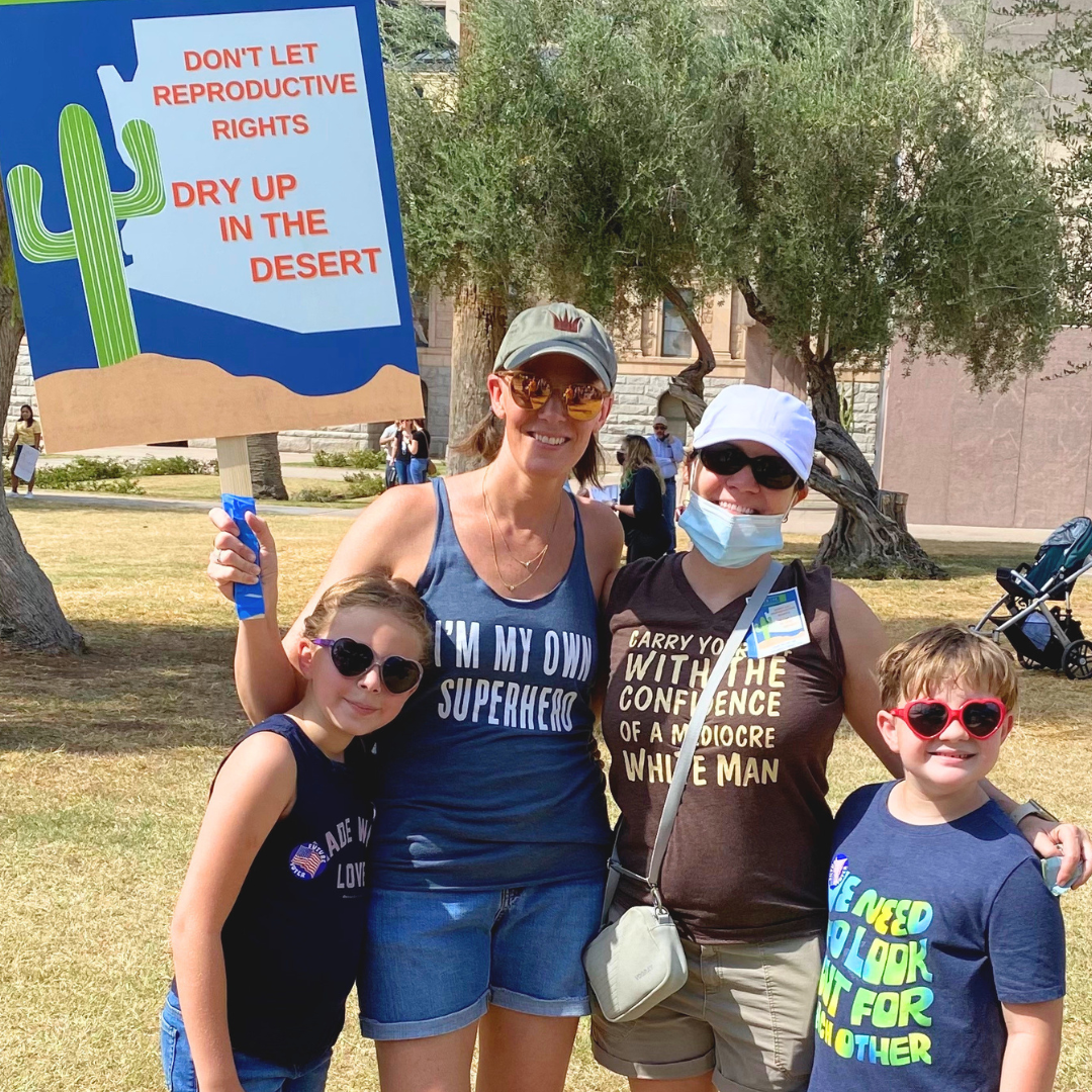 Two women and two children outdoors in a grassy park, smiling, with one holding a protest sign that says "Don't let reproductive rights dry up in the desert," wearing sunglasses and casual clothing.