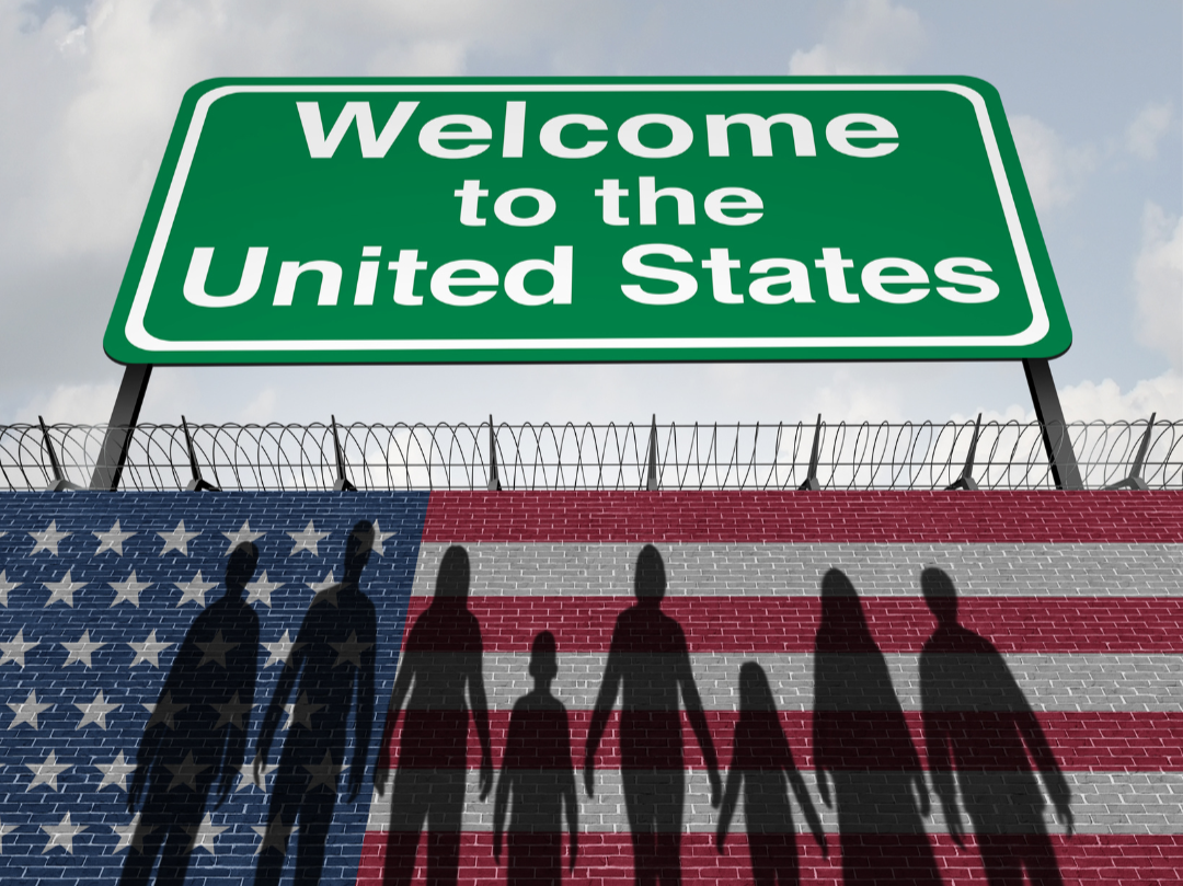 Silhouettes of a diverse group of people holding hands against a brick wall painted with the American flag, with a barbed wire fence on top, underneath a green sign that says "Welcome to the United States" with a cloudy sky in the background.