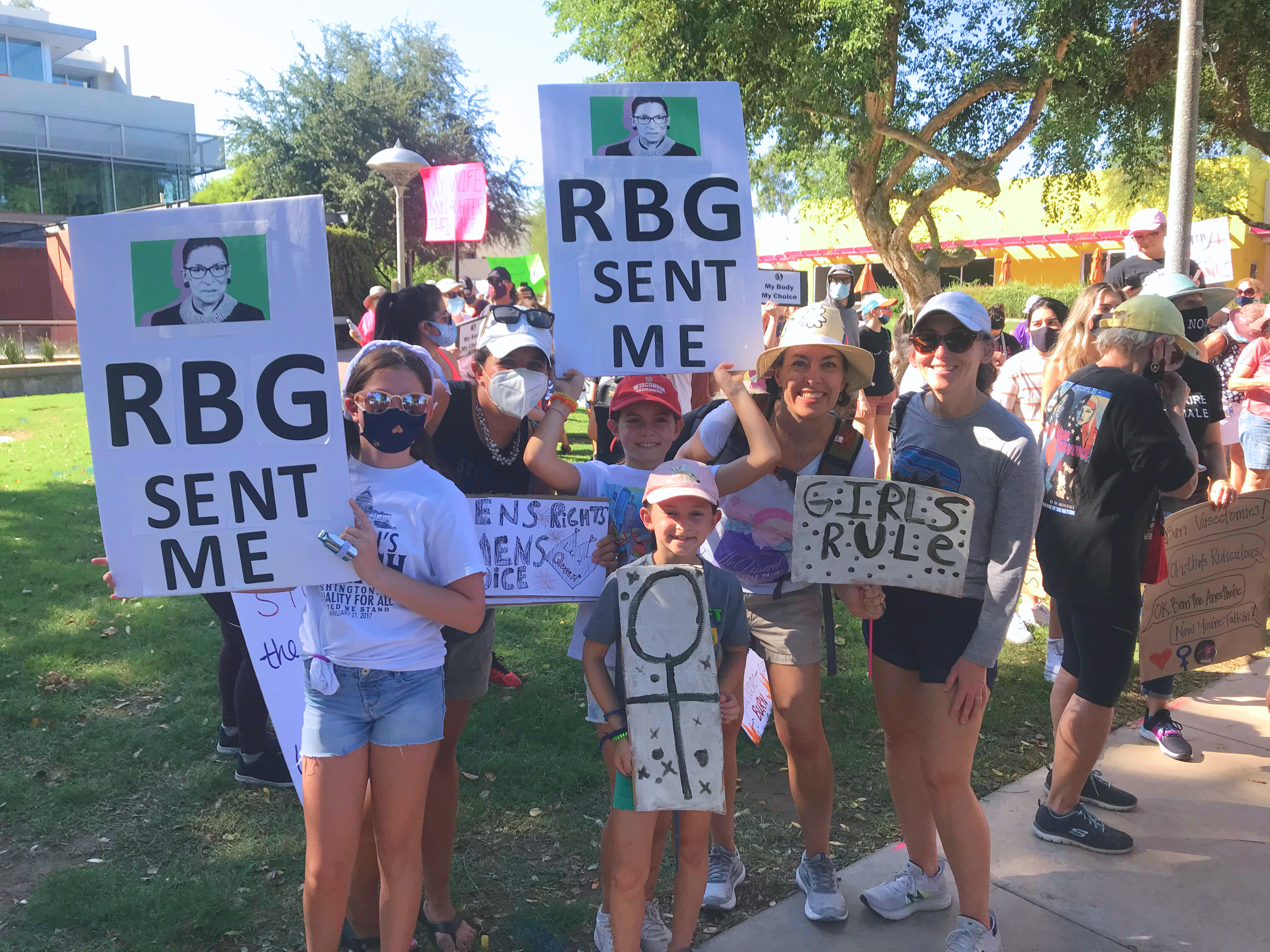 A group of people protesting outdoors, holding signs supporting reproductive rights and justice. Signs include messages like 'RBG Sent Me,' 'Girls R ULE,' and others related to women's rights. Many protesters are wearing masks, and some are wearing hats and sunglasses. There are trees and modern buildings in the background.