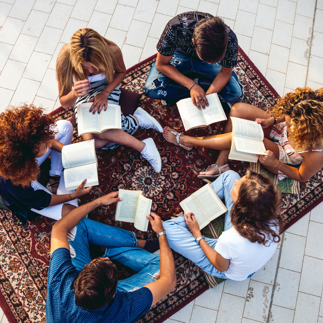 Group of six young people sitting in a circle on a patterned rug, reading books together.