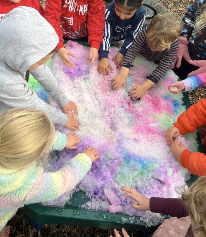 Children playing with colorful synthetic snow or slime at a table outdoors.