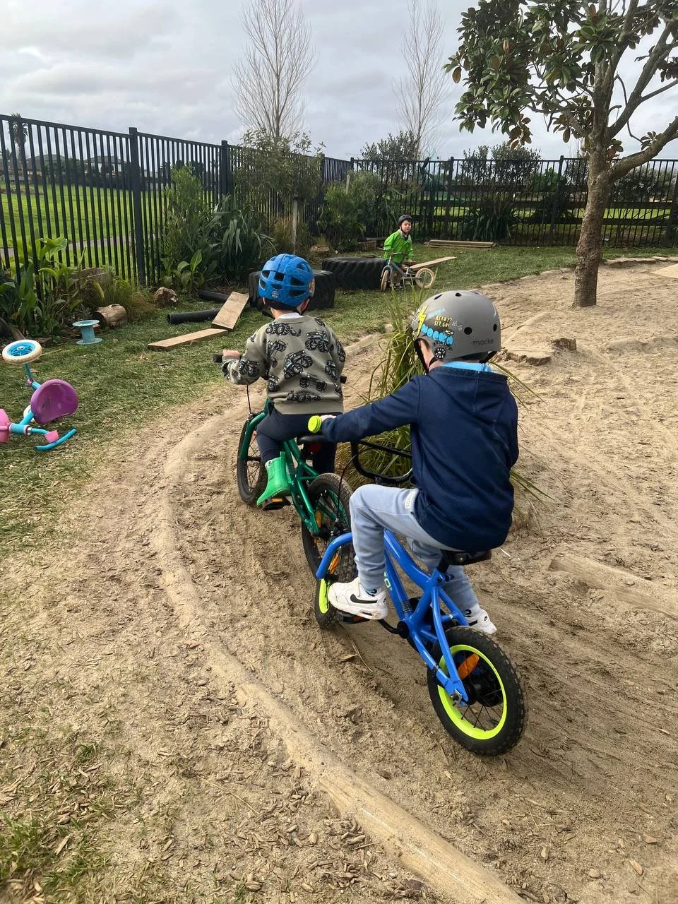 Two children riding bikes on a dirt trail in a playground with a border fence, trees, and grass, while a third child stands in the background.