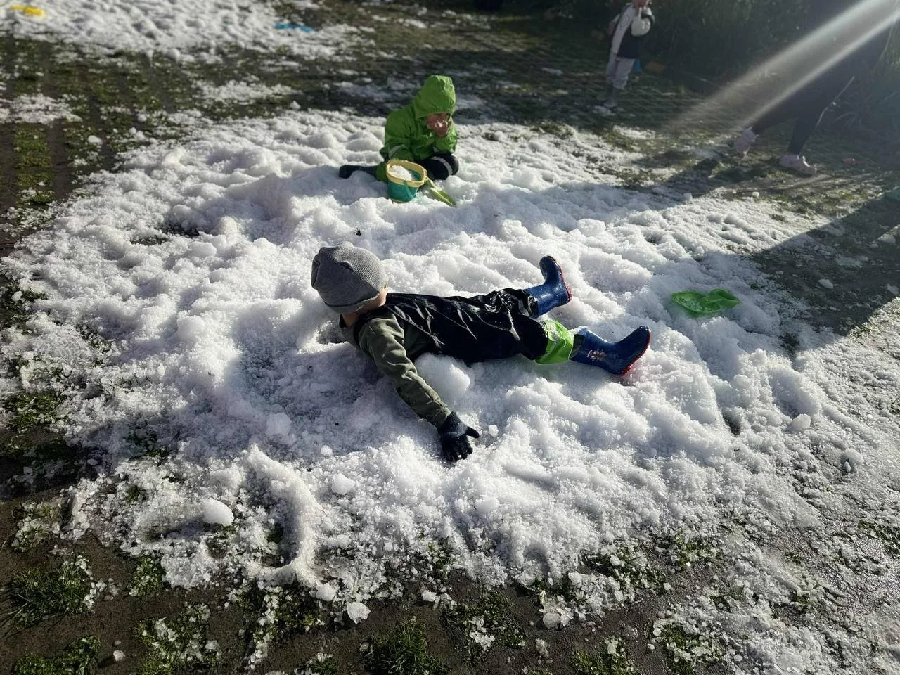 Two children playing in the snow, lying down and surrounded by patches of snow on the ground, with a partially sunny background.