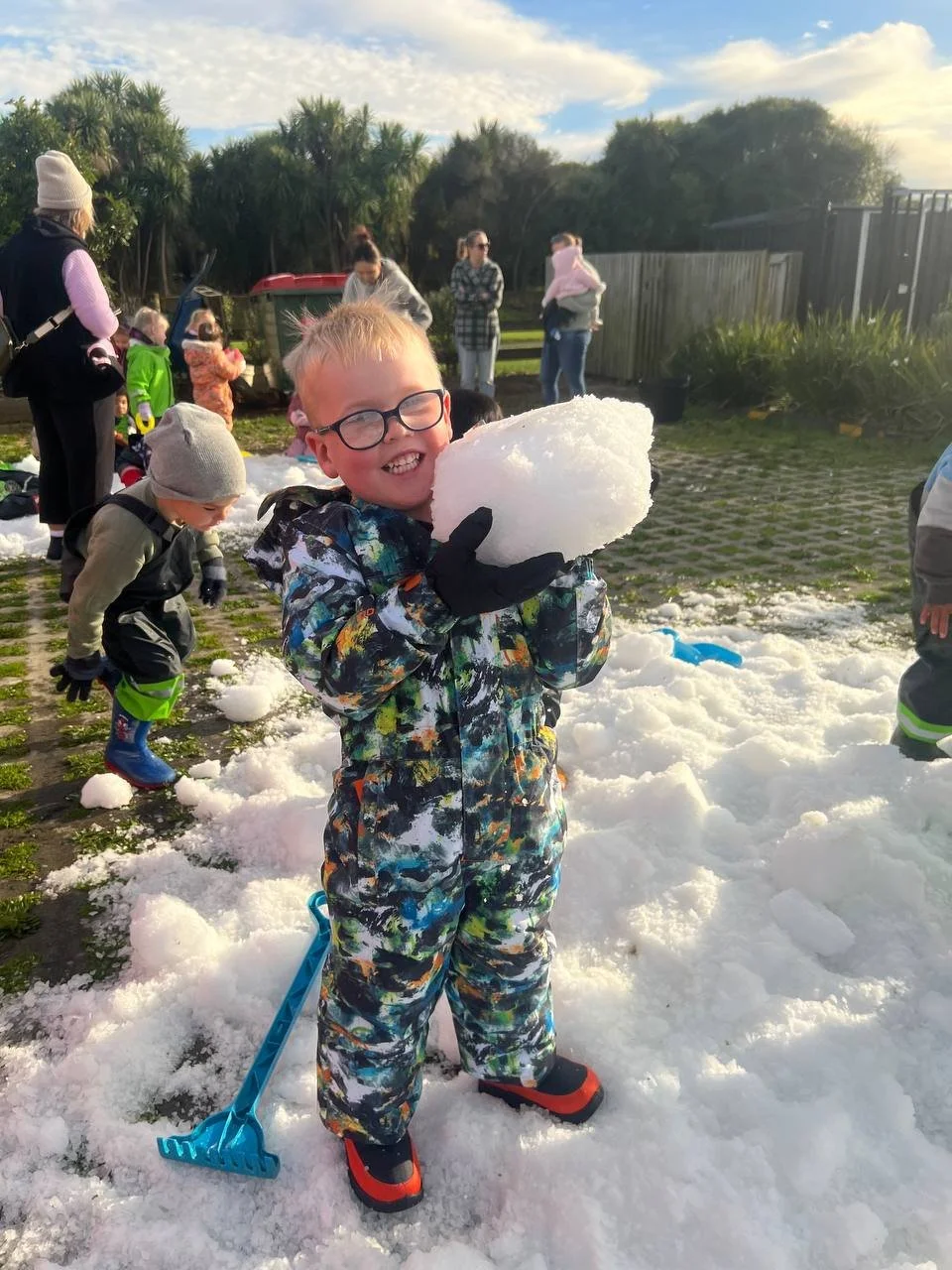 Young boy in colorful winter jacket and boots holding a large snowball, smiling, with others playing in the snow behind him outdoors.