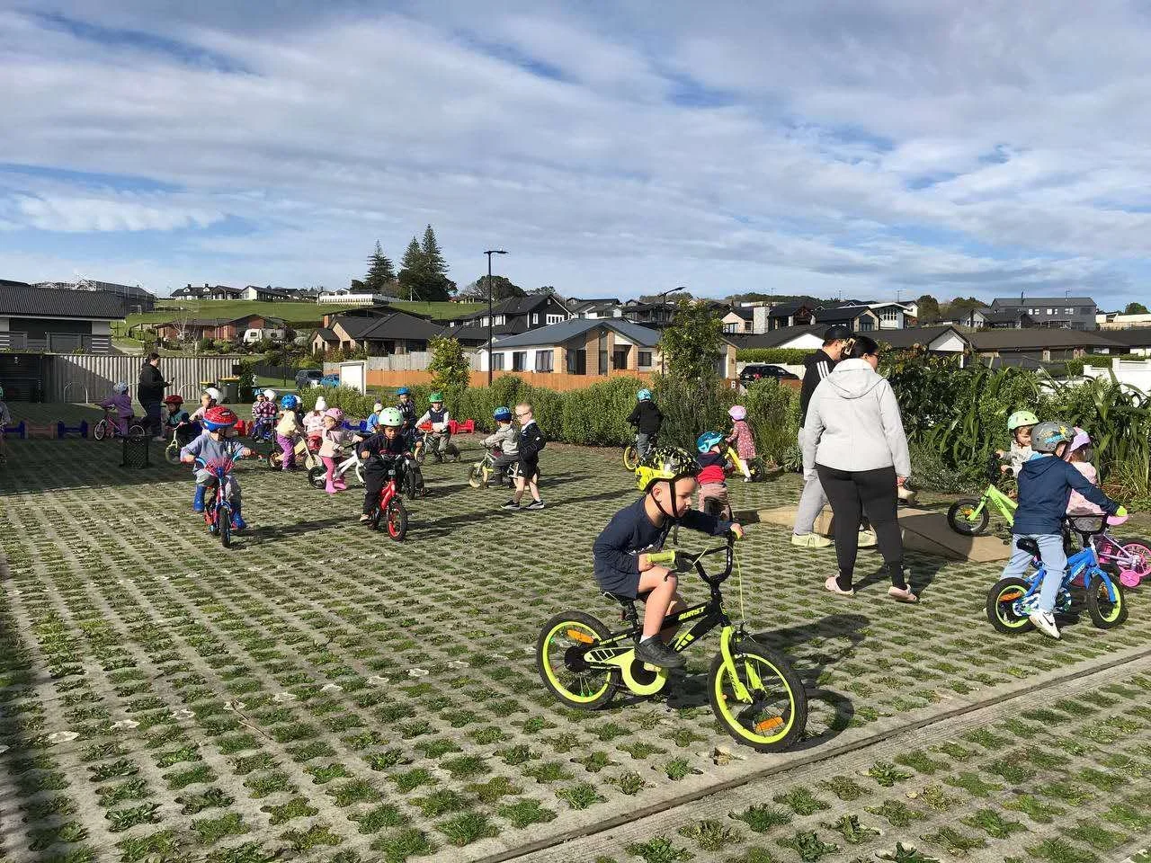 Children riding bikes and learning to ride with supervision in an outdoor parking lot area on a partly cloudy day.