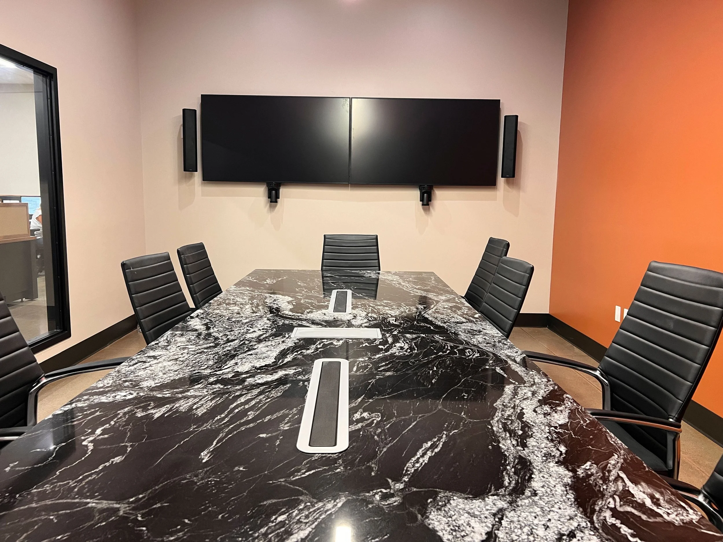 A modern conference room with a black marble table and black leather chairs, a large wall-mounted TV, and two black speakers on either side of the TV, with a window on the left and a multicolored accent wall on the right.