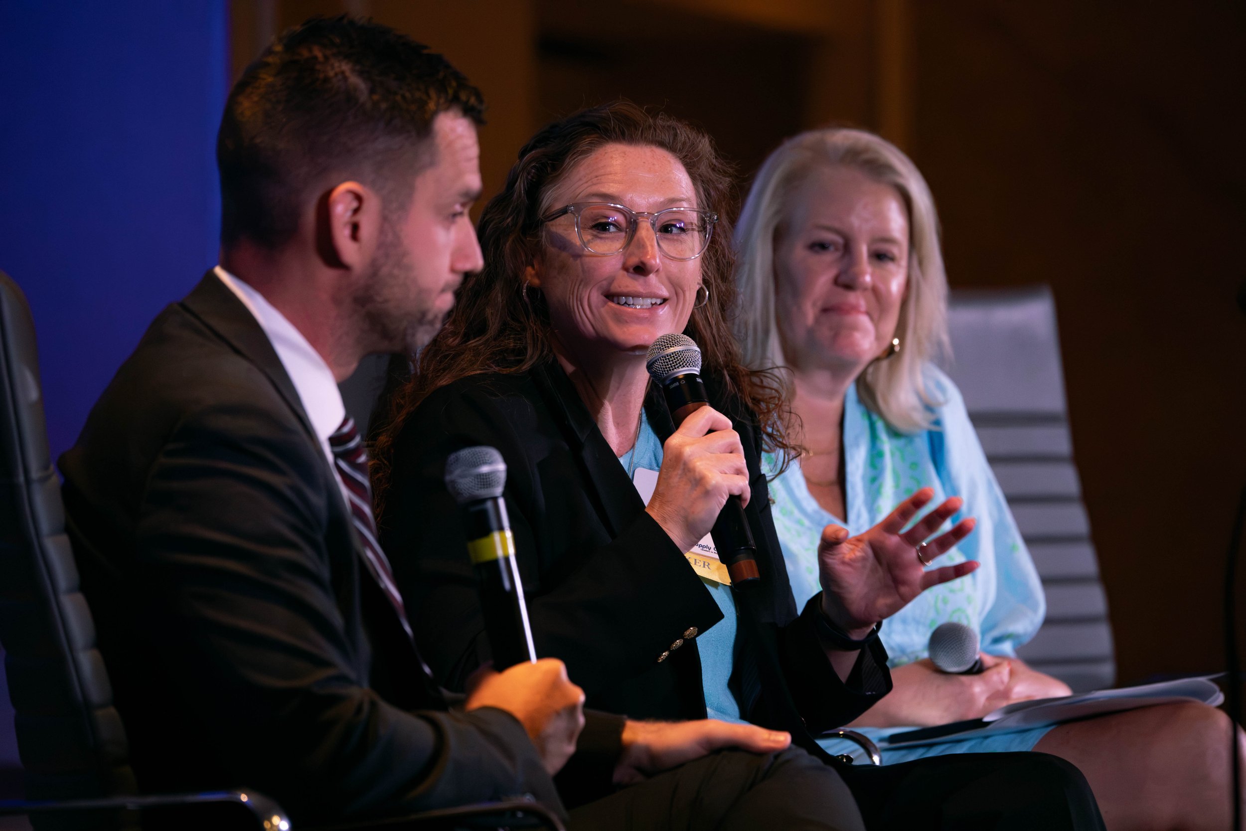 A panel of four people sitting at a table during a discussion or conference, with one woman holding a microphone and speaking while others listen.
