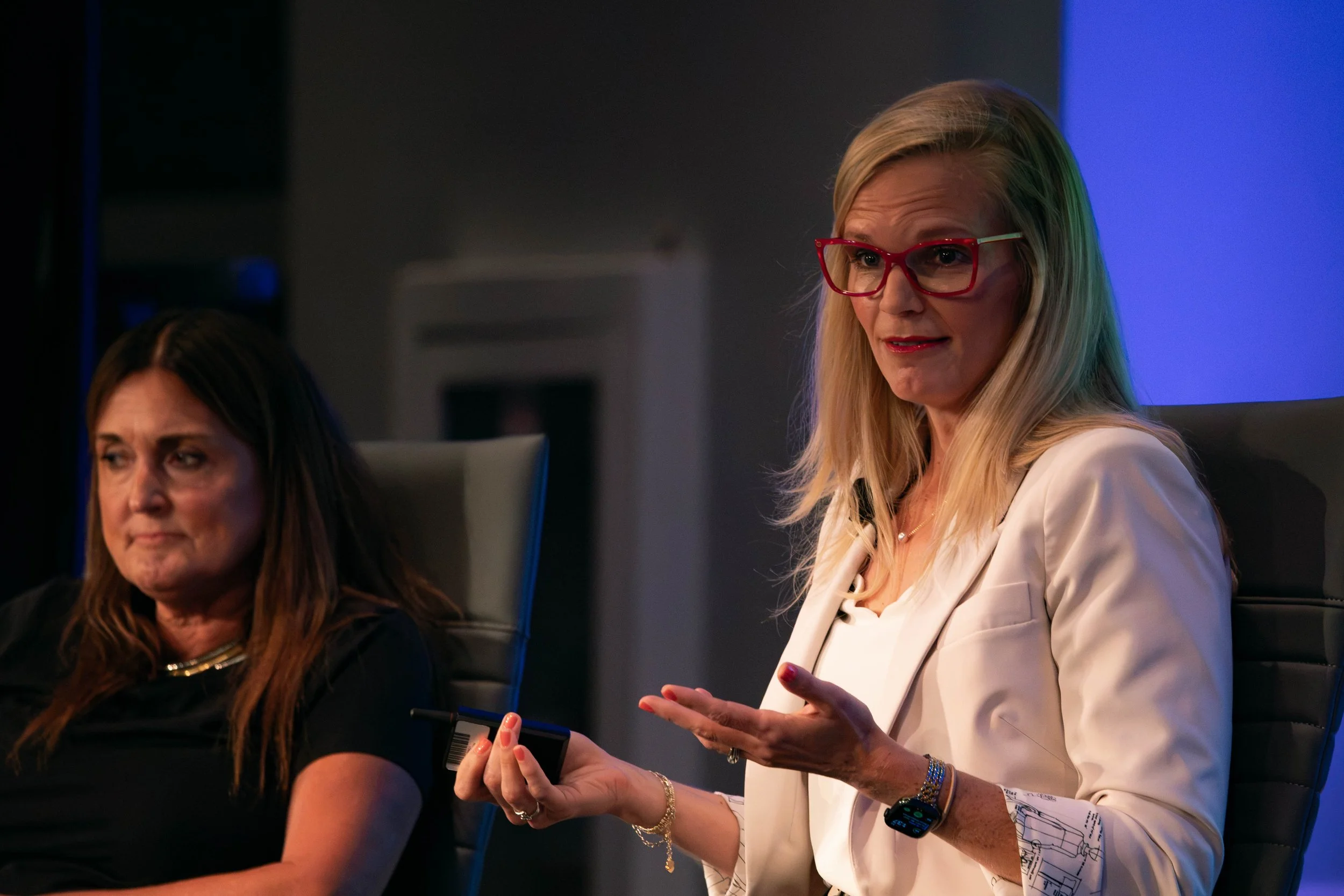 Two women sitting at a table during a meeting or presentation, one woman with long blonde hair and red glasses speaking, holding a remote or presentation device, the other with long dark hair listening.