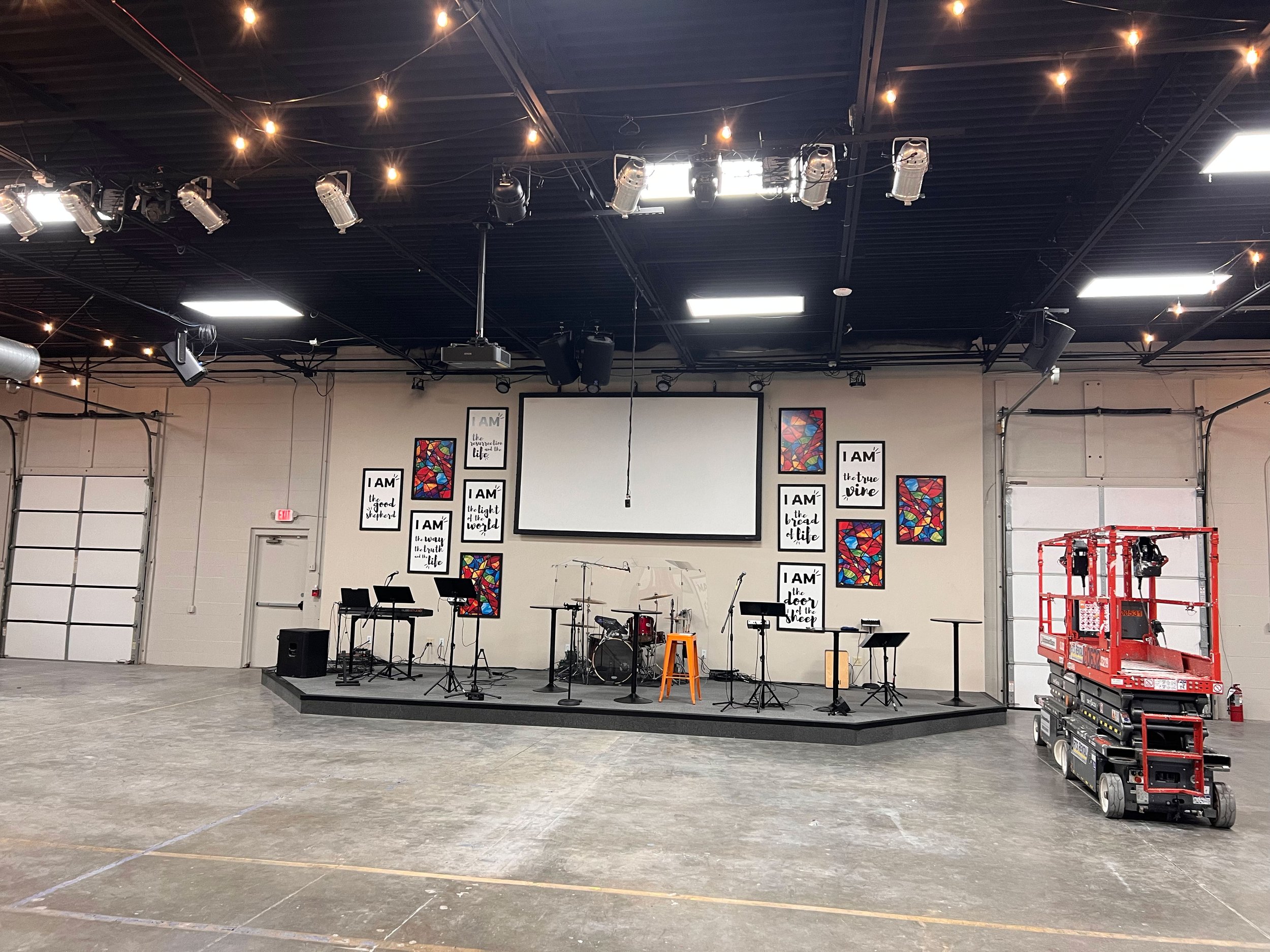 An indoor stage setup with musical instruments like drums, keyboard, and microphones, surrounded by a large open space with a concrete floor, framed artworks on the wall, ceiling lighting, and equipment like a cherry picker and speakers.