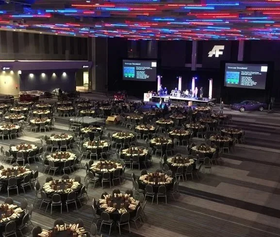 Ballroom set up with round tables and chairs, stage with a band or speakers, and a large screen displaying event information