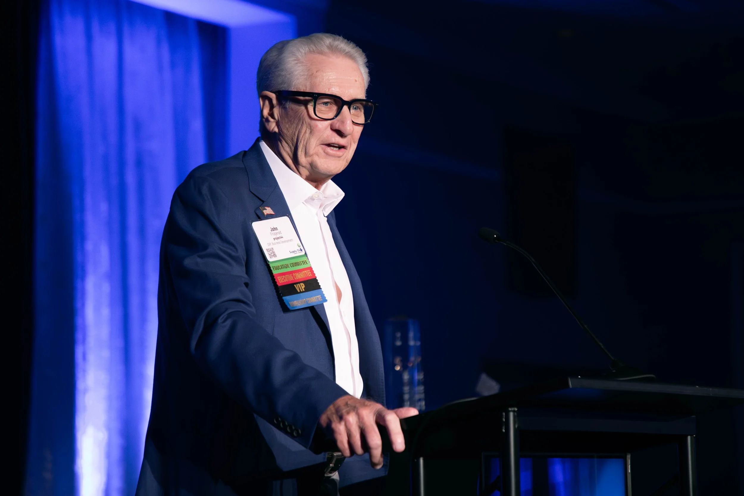 An elderly man with gray hair and glasses speaking at a podium during a conference or event. He is wearing a navy blue suit, white shirt, and a name badge with various colorful ribbons. The background features blue curtains and low lighting.