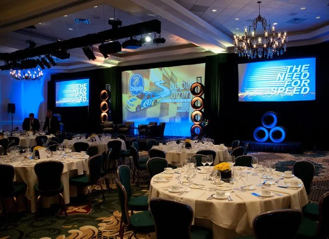 Elegant banquet hall with round tables set for dinner, decorated with yellow floral centerpieces, and a stage with large screens displaying 'The Need for Speed' logo and racing imagery, chandeliers hanging from ceiling.