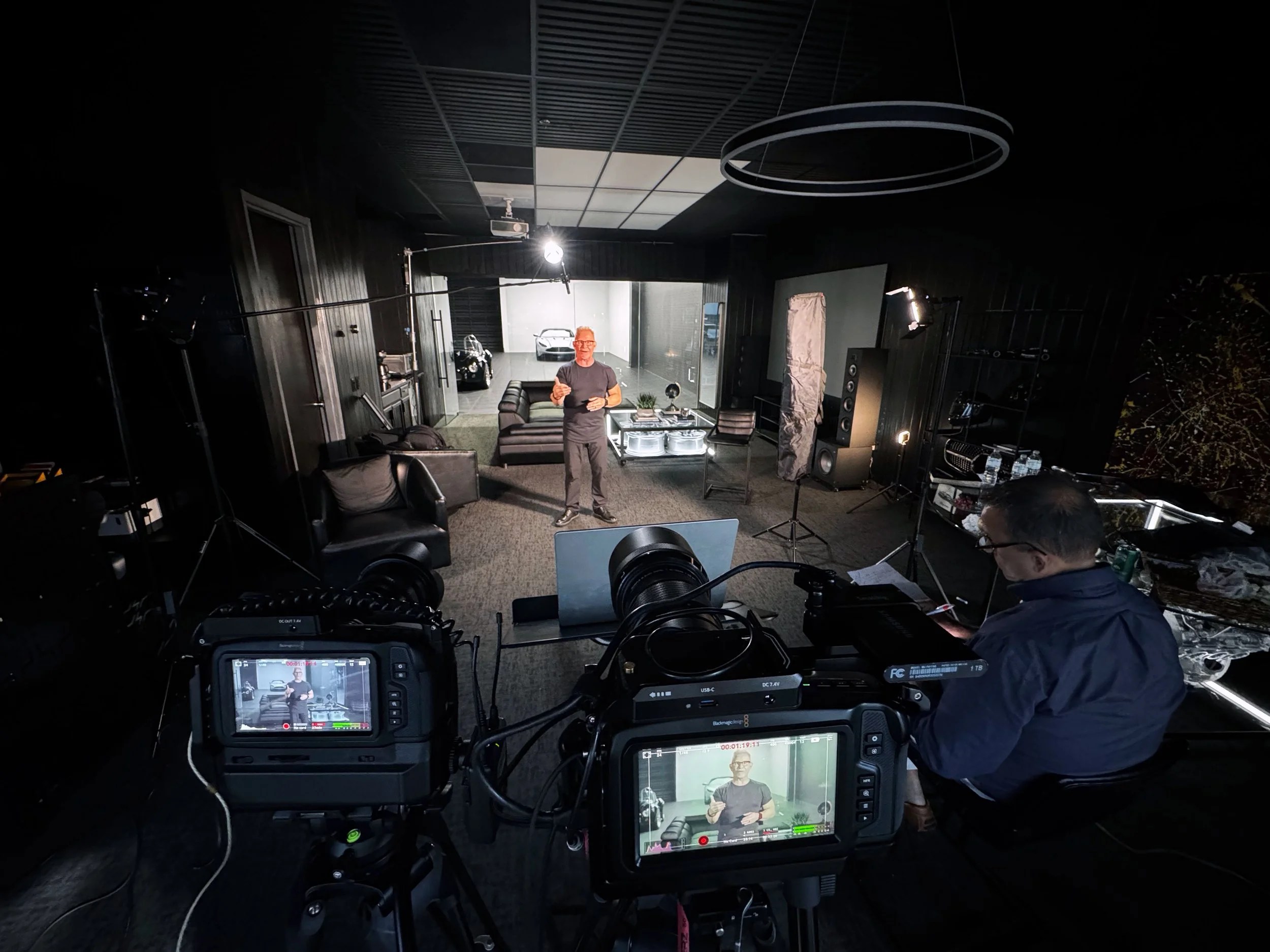 A man in a grey t-shirt and glasses standing in a room with black walls and ceiling, speaking in front of a camera setup. There are sofas, a table with a small plant, speakers, and a large screen showing the man.