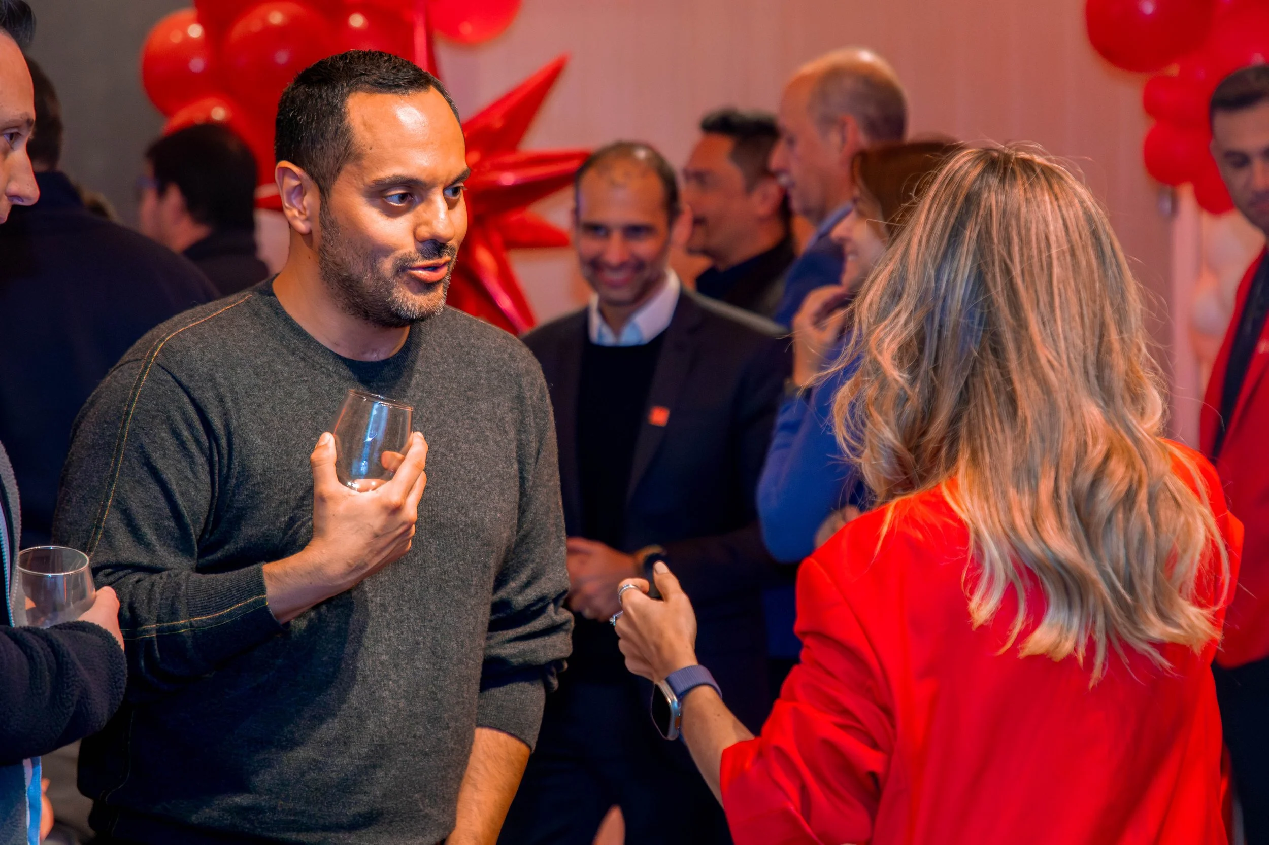 Group of people at a social event, including a man in a gray sweater holding a wine glass greeting a woman in a red jacket.