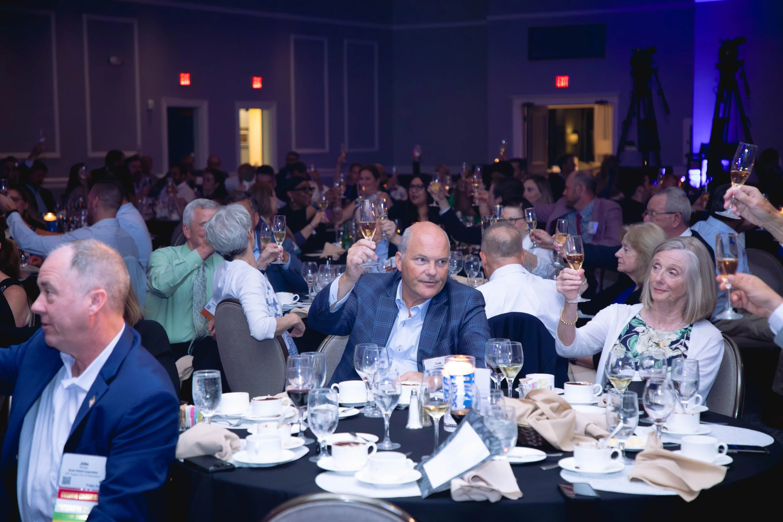 A large group of people seated at round tables in a conference or banquet hall, raising glasses of champagne in a toast, with some people smiling and engaging in conversation, decorated with table settings and cups, with blue and dim lighting and large screen monitors on the walls.