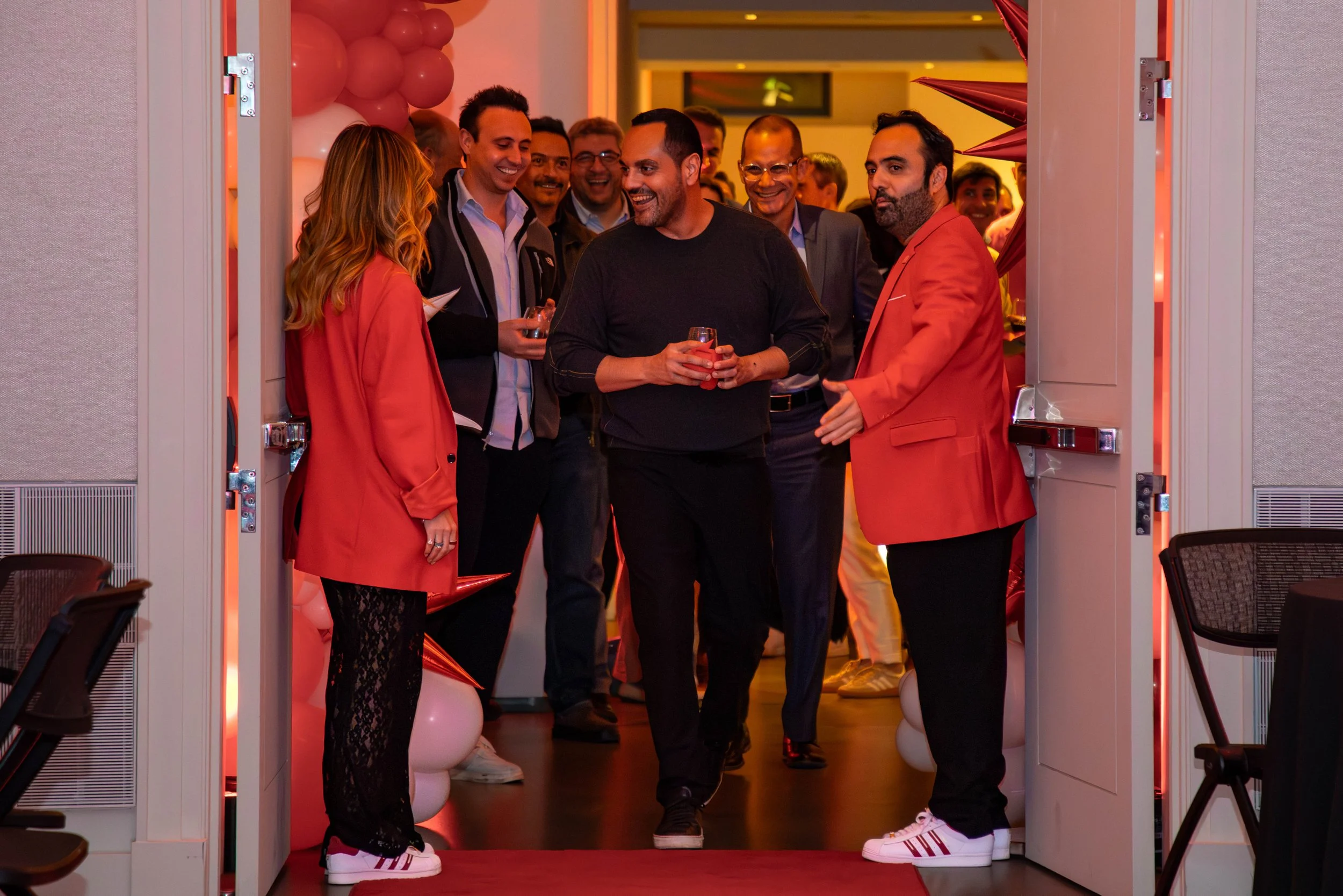A group of people smiling and talking at an event, standing in a doorway decorated with balloons, with some holding drinks.
