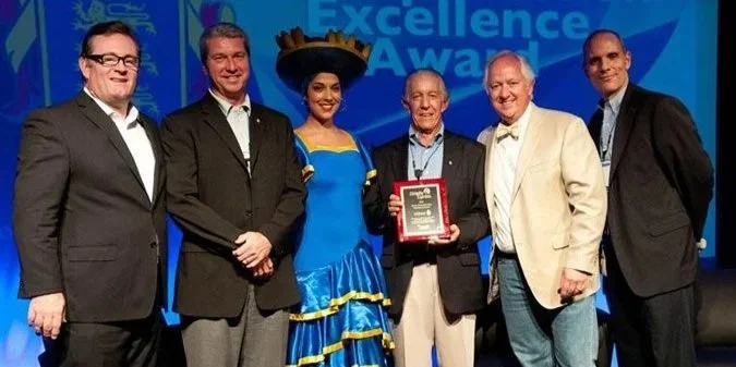 Six people standing together on a stage at an awards ceremony, with a screen in the background displaying "Excellence Awards." One person in the center holds a plaque, and another is dressed in a traditional blue and yellow dress with a large hat.