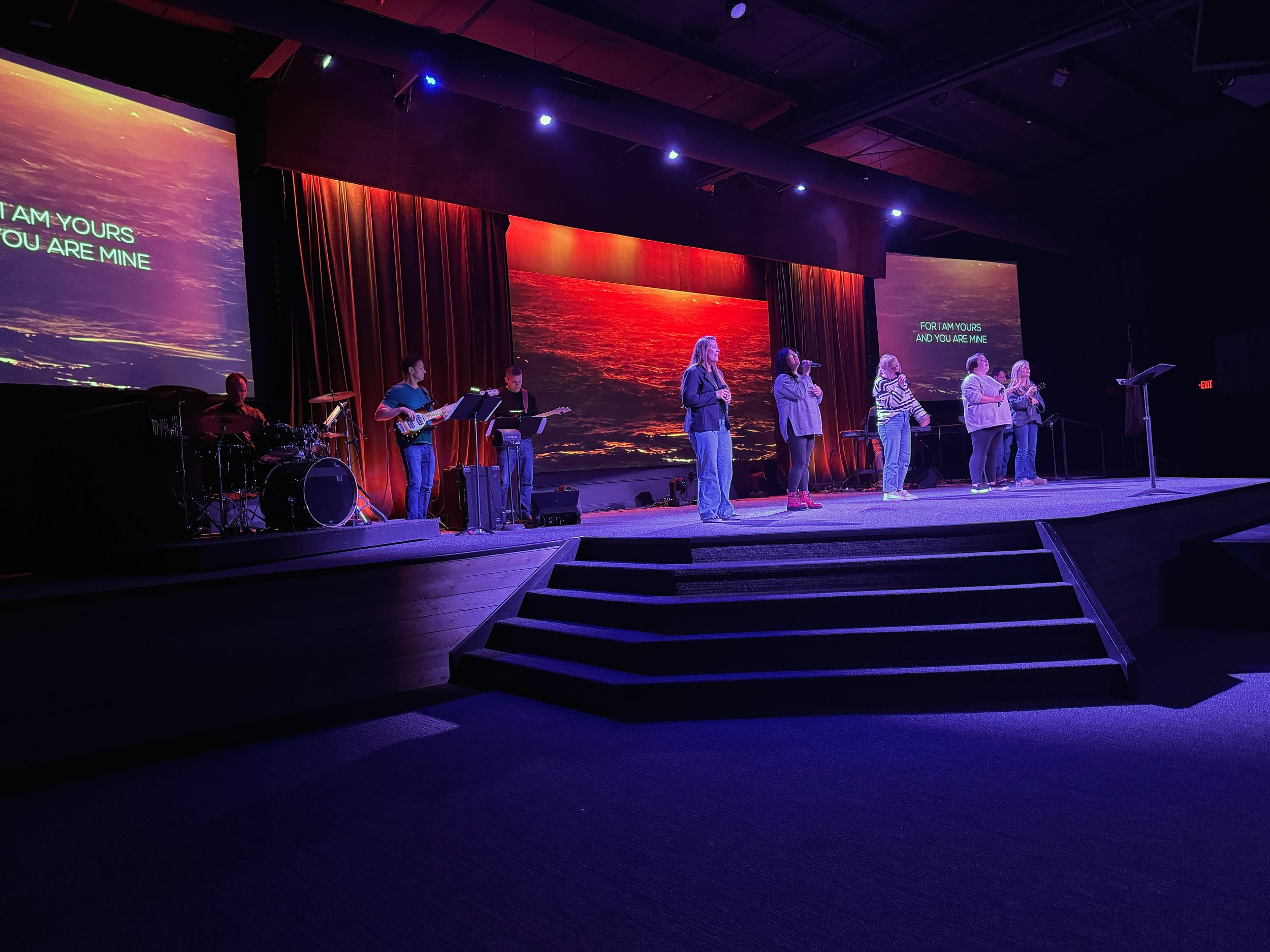 Stage with musicians and large screens displaying ocean sunset and lyrics, dim lighting, auditorium setting.