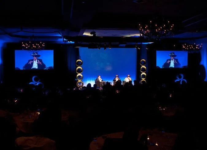 A darkened conference or event room with a stage, featuring three people seated and speaking. The stage has large circular decorations and is illuminated with blue lighting. Two large screens on either side of the stage display a close-up of one speaker. The room has chandeliers hanging from the ceiling and an audience seated at tables.