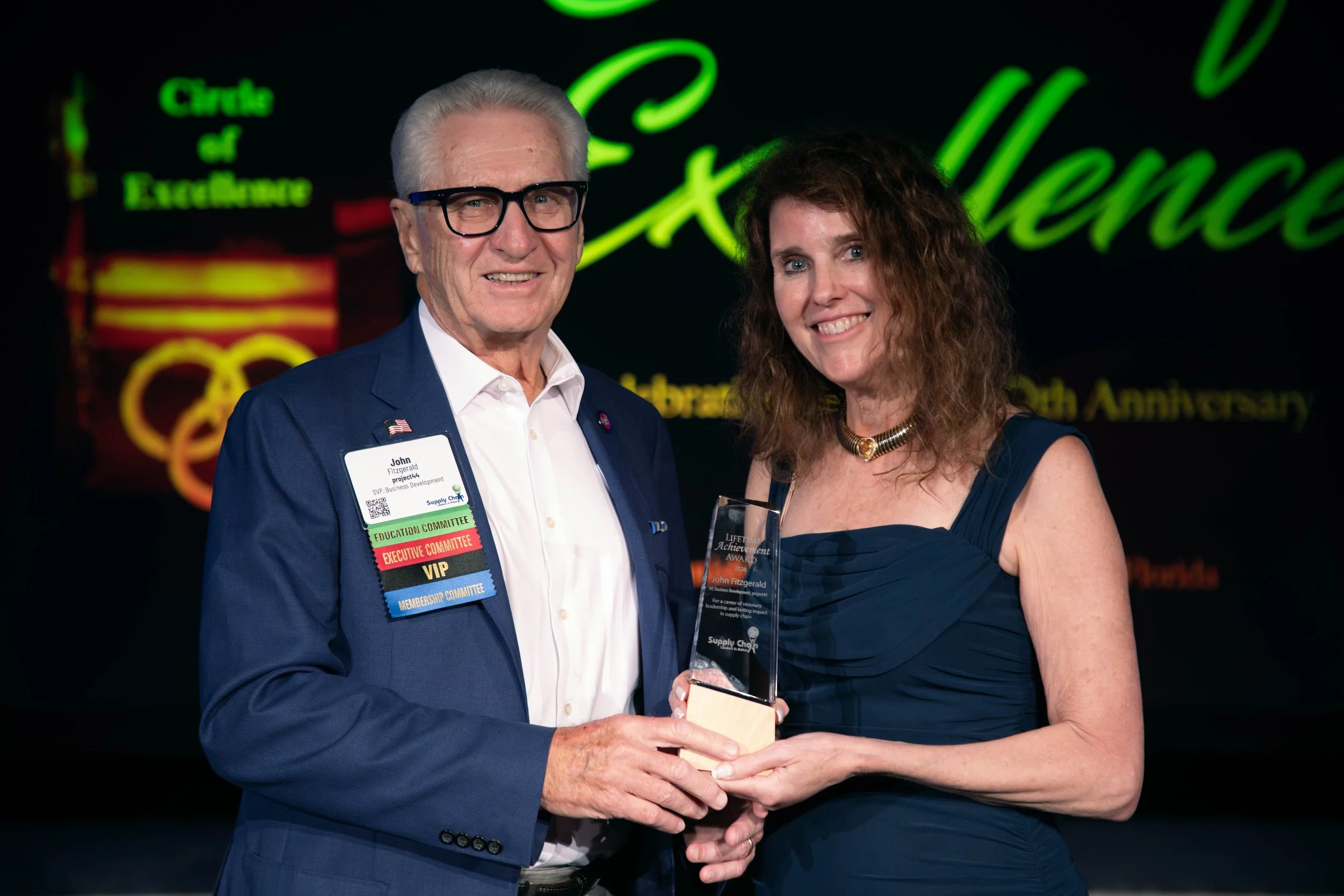 A man in a blue suit and woman in a dark blue dress are on stage, smiling. The man is holding an award, and the woman is receiving it. The background features a neon sign with the words 'Circle of Excellence' and 'Anniversary'.