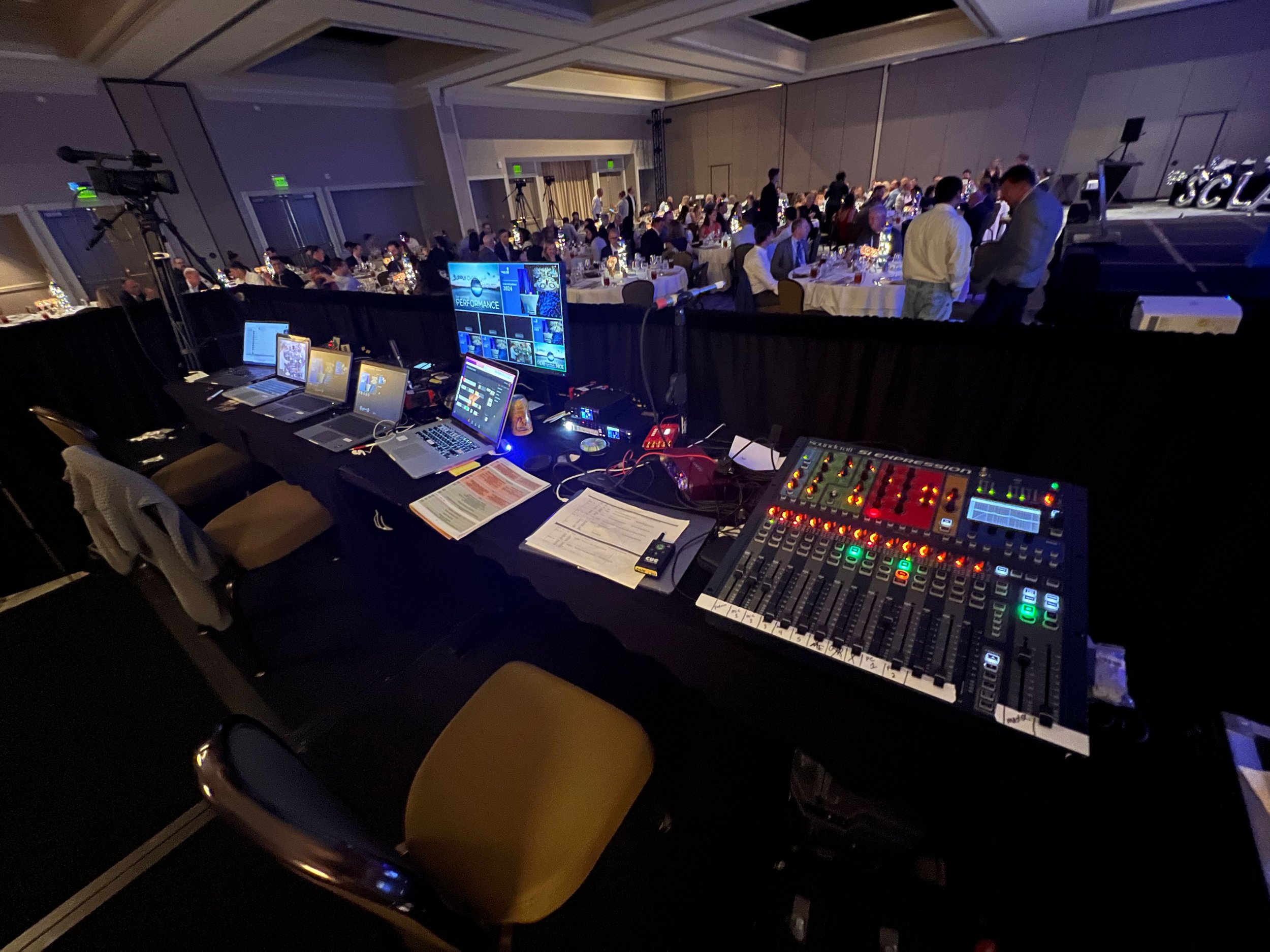 View of a large conference room filled with round tables and many people seated, decorated with blue lighting. In the foreground, there is technical equipment including multiple laptops, computers, and a sound mixing console on a table. The room has a stage area with large decorative letters and a screen, indicating a professional event or conference.