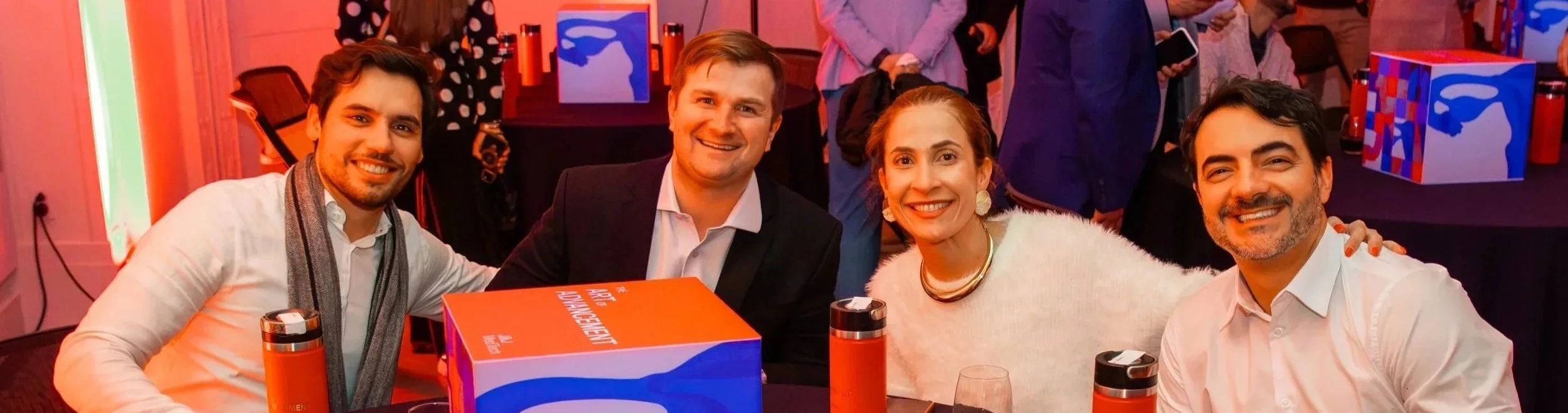 Four smiling adults sitting at a table during a celebration event, with colorful decorations and gift boxes in the background.