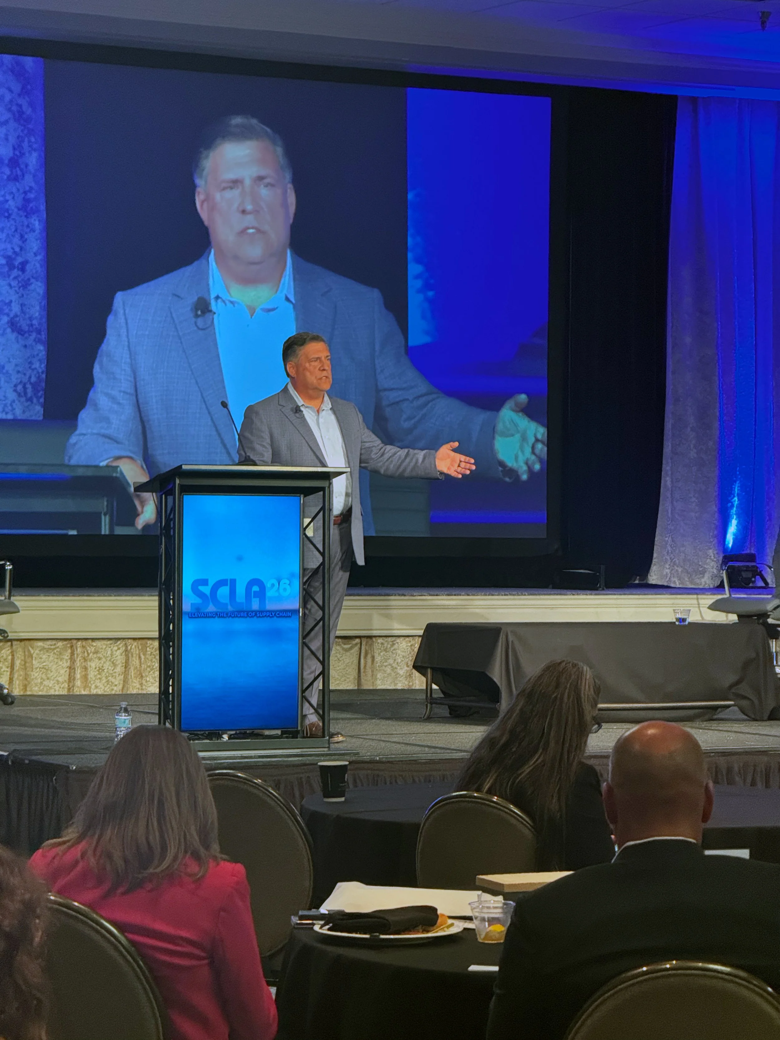 A man in a gray suit speaks at a conference with a lectern displaying a blue and white logo. A large projection screen behind him shows his image. The audience is seated at round tables with black tablecloths.