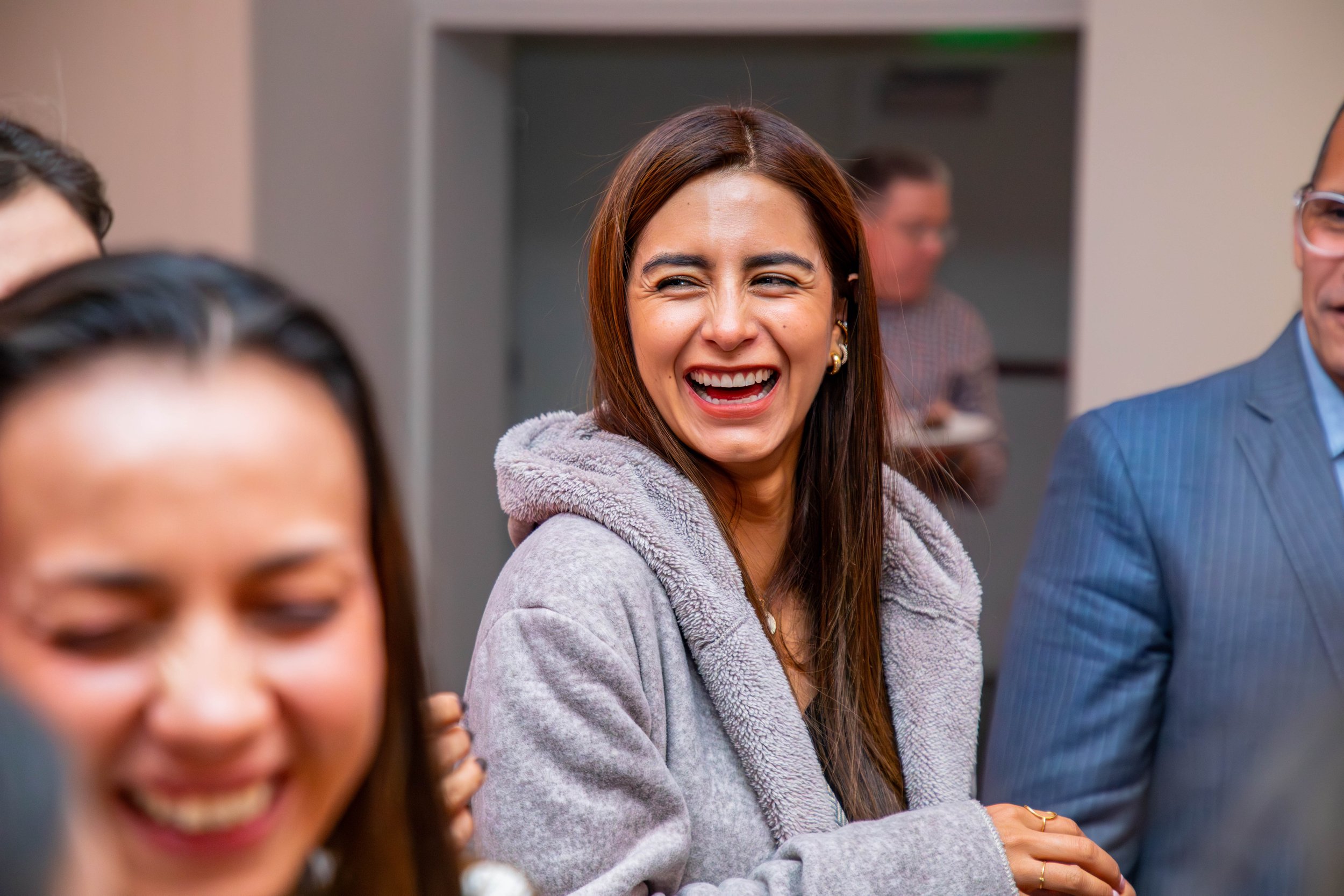 A woman with long brown hair, smiling and wearing a gray hoodie, is laughing in a crowd of people.
