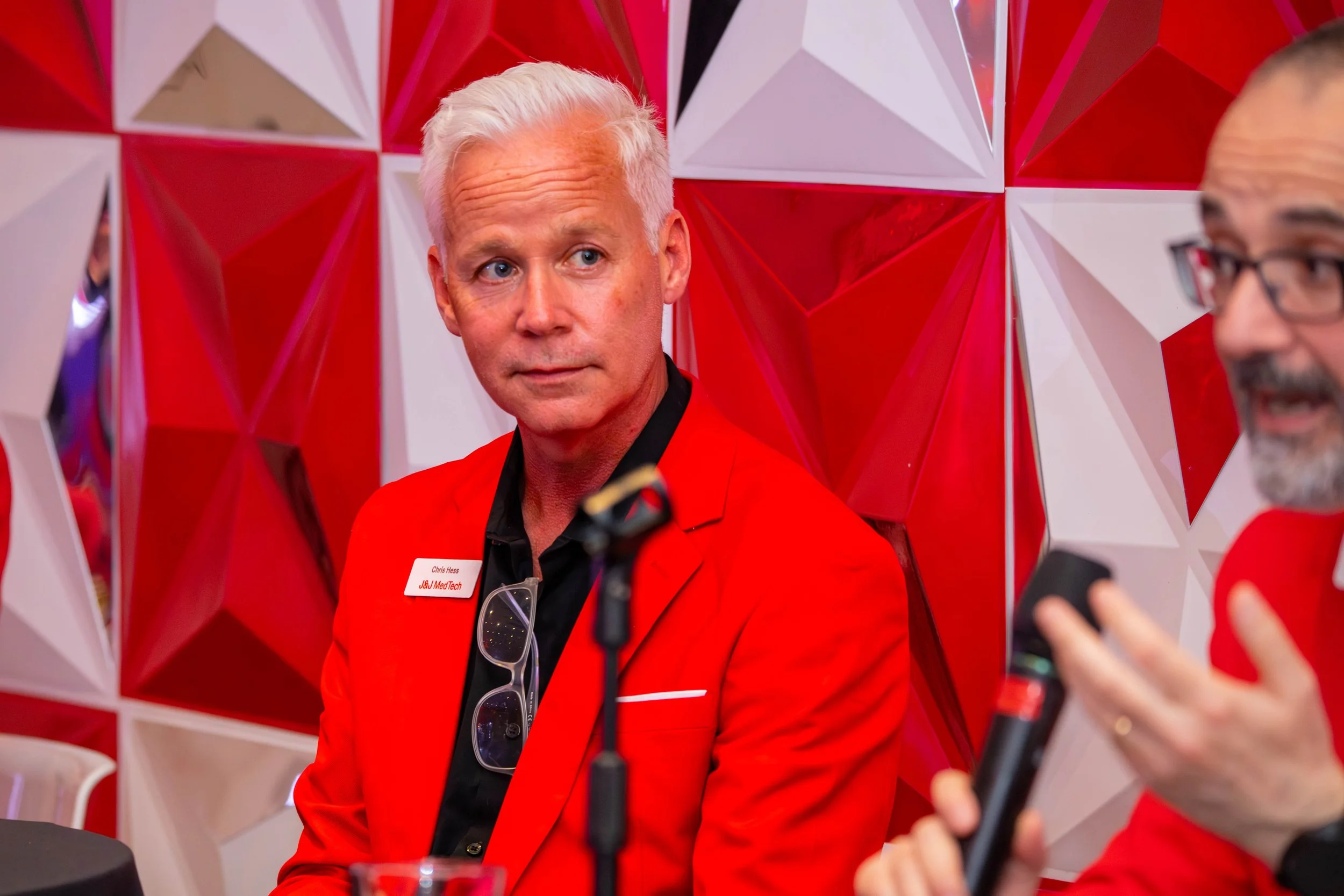A man with white hair wearing a red jacket and black shirt, sitting at a table at a press conference or interview, with a geometric red and white background behind him.
