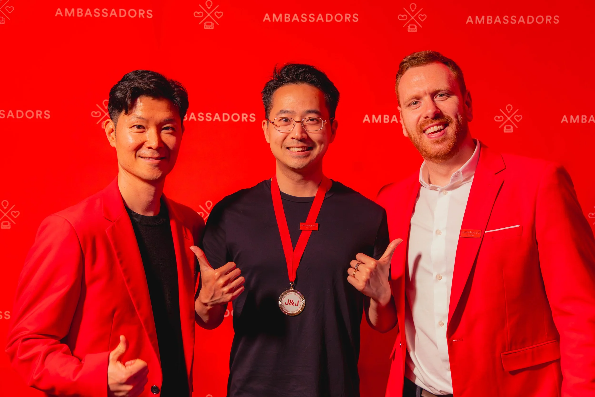 Three men in red blazers standing in front of a red backdrop with the word "AMBASSADORS" repeated. The man in the middle is holding a medal with the initials J&J and showing a thumbs-up while smiling.