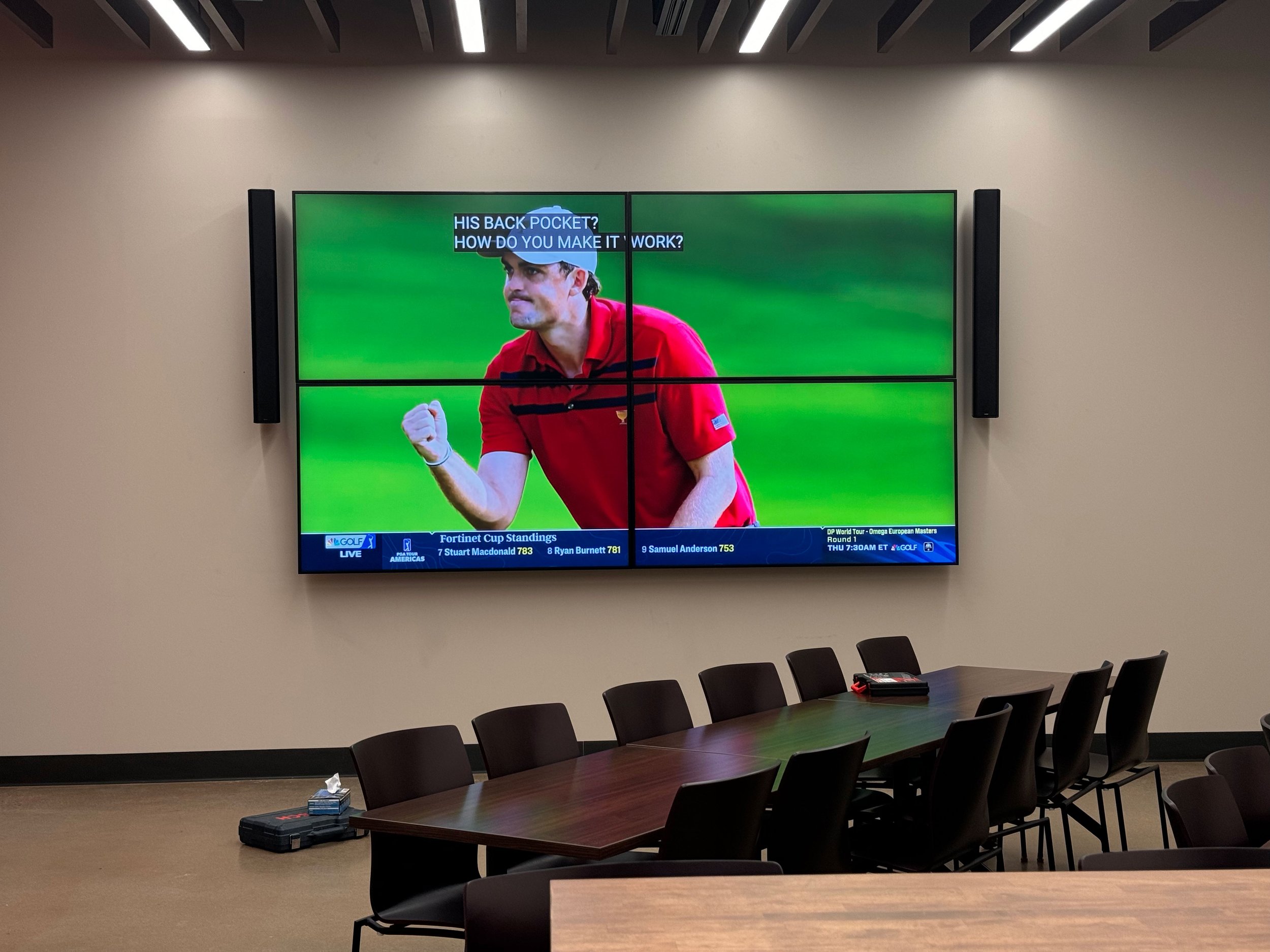 A conference room with a long table and black chairs, and a large wall-mounted TV screen showing a golfer in a red shirt. The room has beige walls and a modern ceiling design.