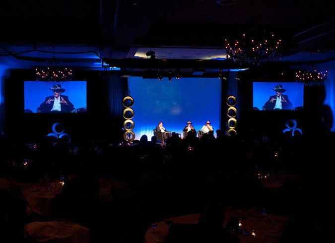 A stage with three people seated and a large screen displaying the image of a person speaking, illuminated with blue lighting, decorated with circular gold accents, chandeliers hanging from the ceiling, and an audience in a darkened banquet hall.