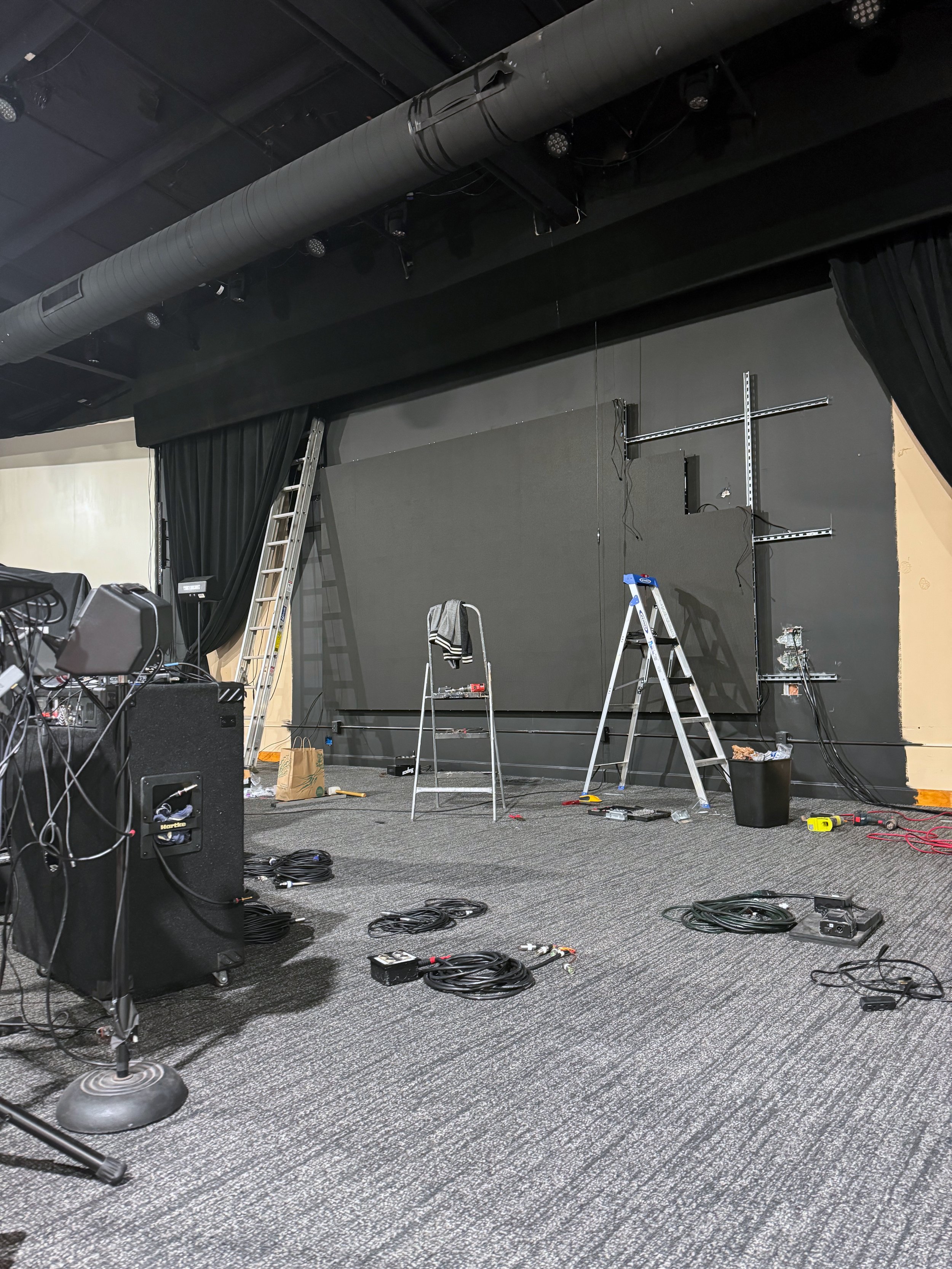 Stage setup with ladders, speakers, cables, and a large screen, indicating preparations for a performance or event.