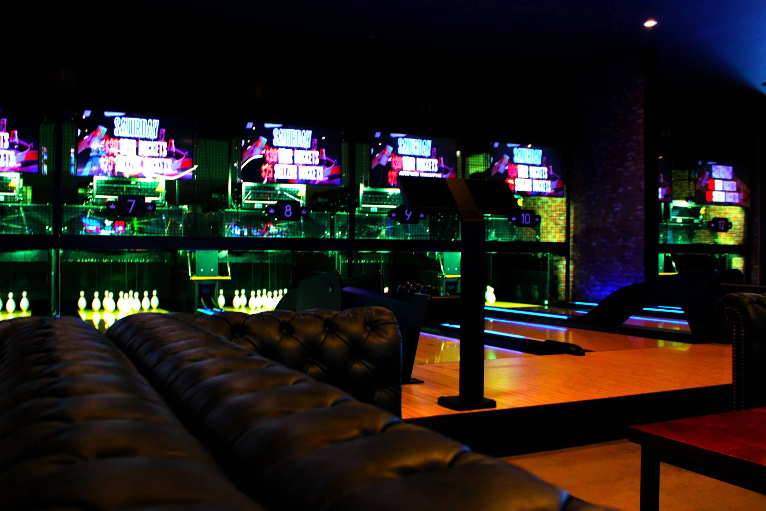 Indoor area with multiple large digital screens displaying promotional content, neon lights, and bowling pins on a shelf, resembling a bowling alley or entertainment venue.