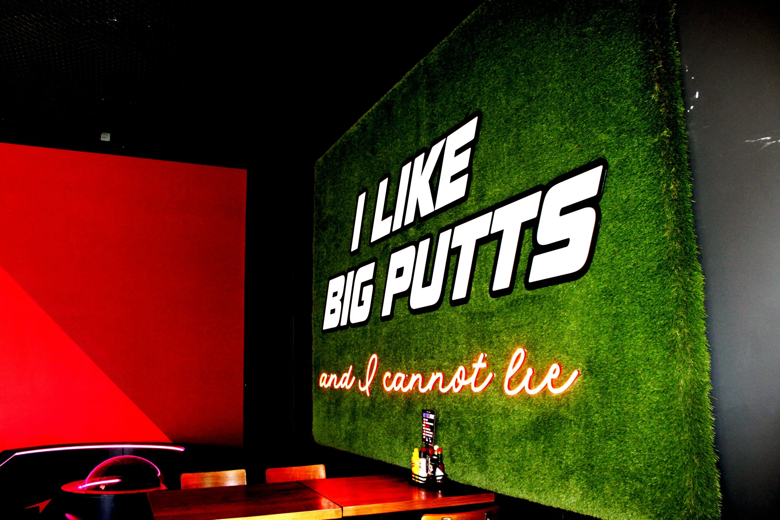 Interior of a restaurant with a green artificial grass wall featuring the bold white logo 'LIKE BIG POUTS'. There is a red neon sign with part of a phrase visible that reads 'and I cannot lie.' A wooden table with condiment bottles and a pink light reflecting off the furniture is in the foreground.