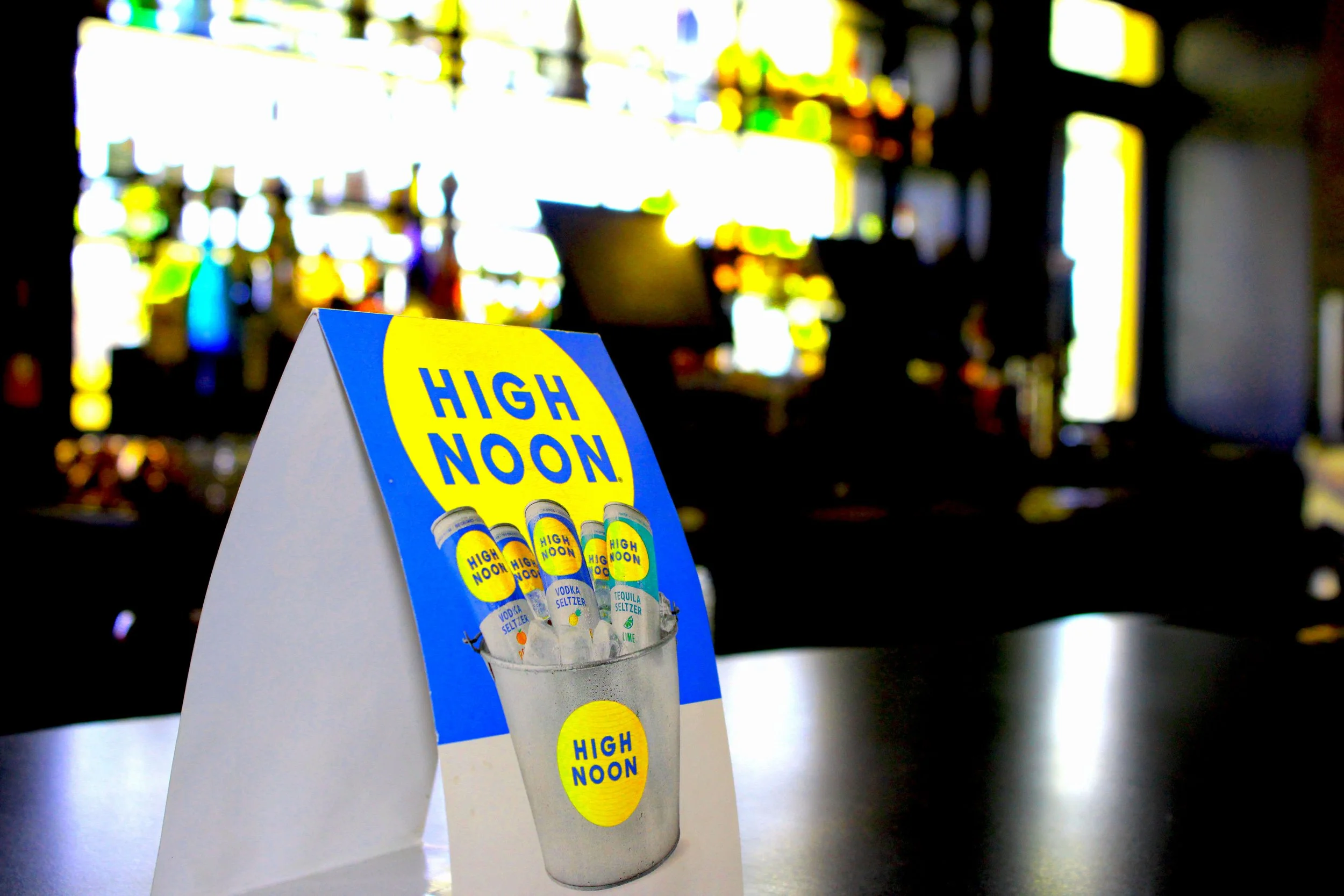 A table tent displaying a selection of flavored vodka seltzers with a blue and yellow high noon branding card behind it, set against a blurred background with bright city lights.