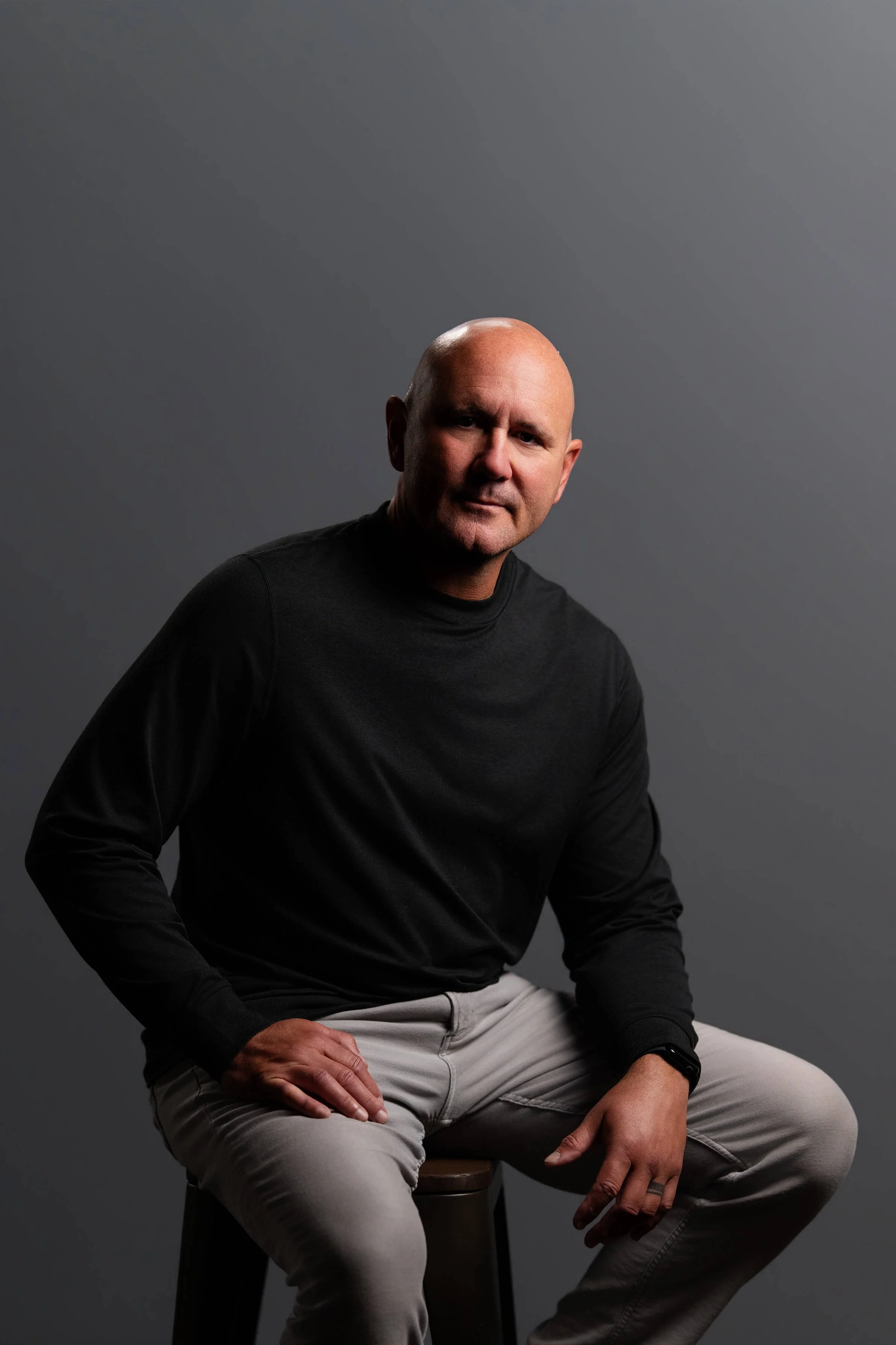 A man with a shaved head wearing a black long-sleeve shirt and light-colored pants, sitting on a stool against a plain gray background.