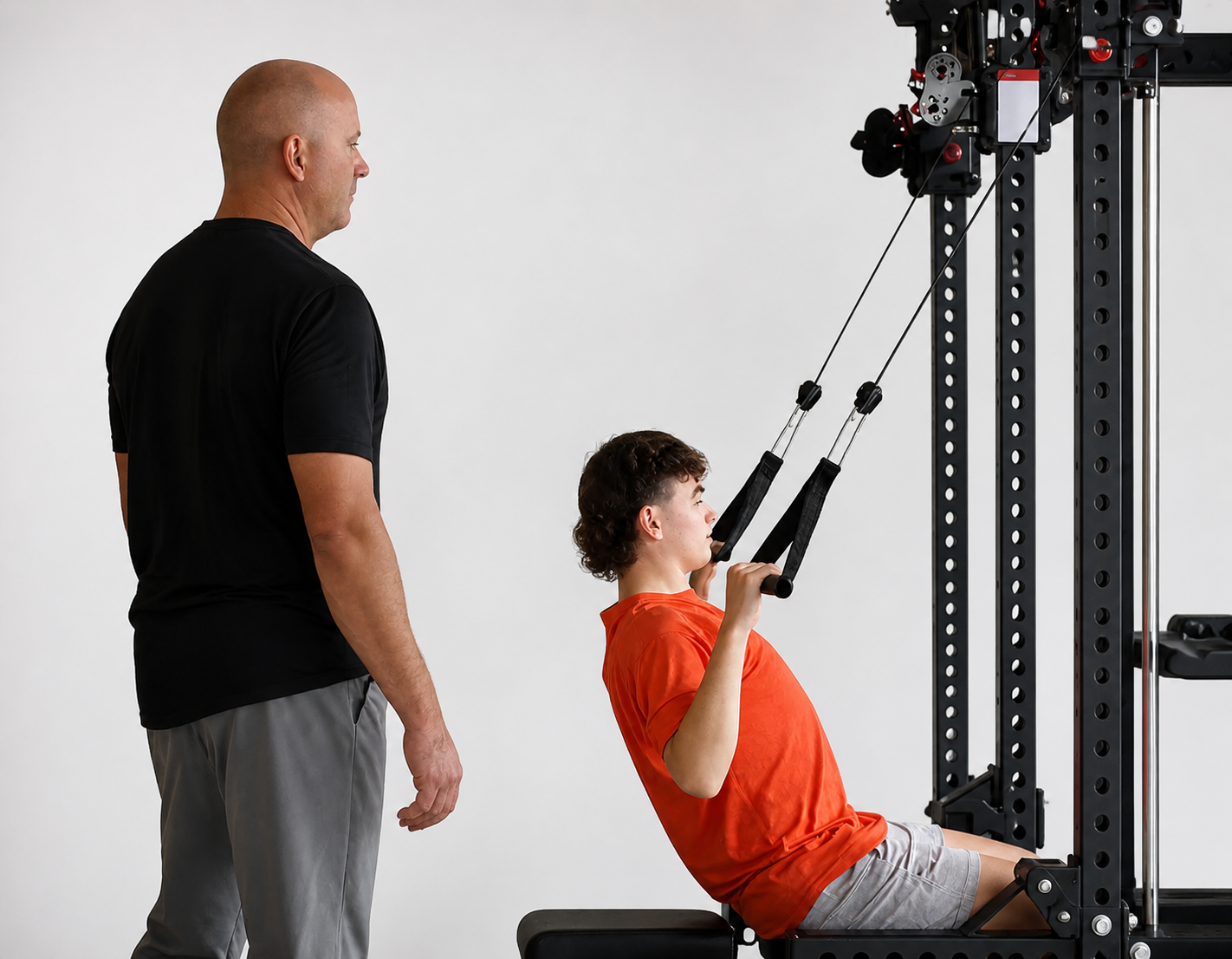 A young man in an orange shirt and gray shorts seated and exercising with a pulley machine, guided by an instructor in a black shirt and gray pants, in a fitness studio with a plain white background.