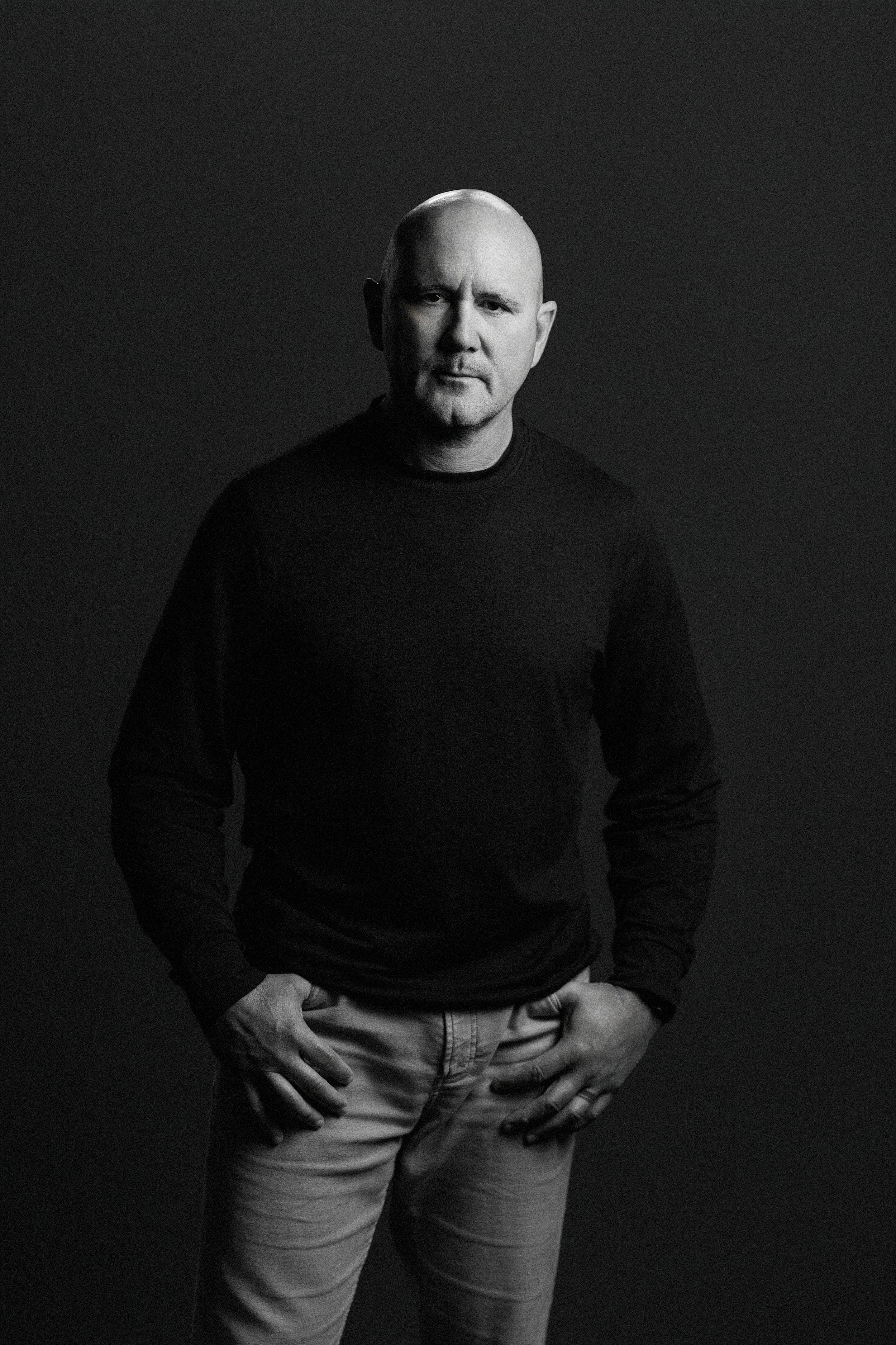 Black and white photograph of a bald man wearing a black long-sleeve shirt and light-colored pants, standing with hands on hips against a dark background.