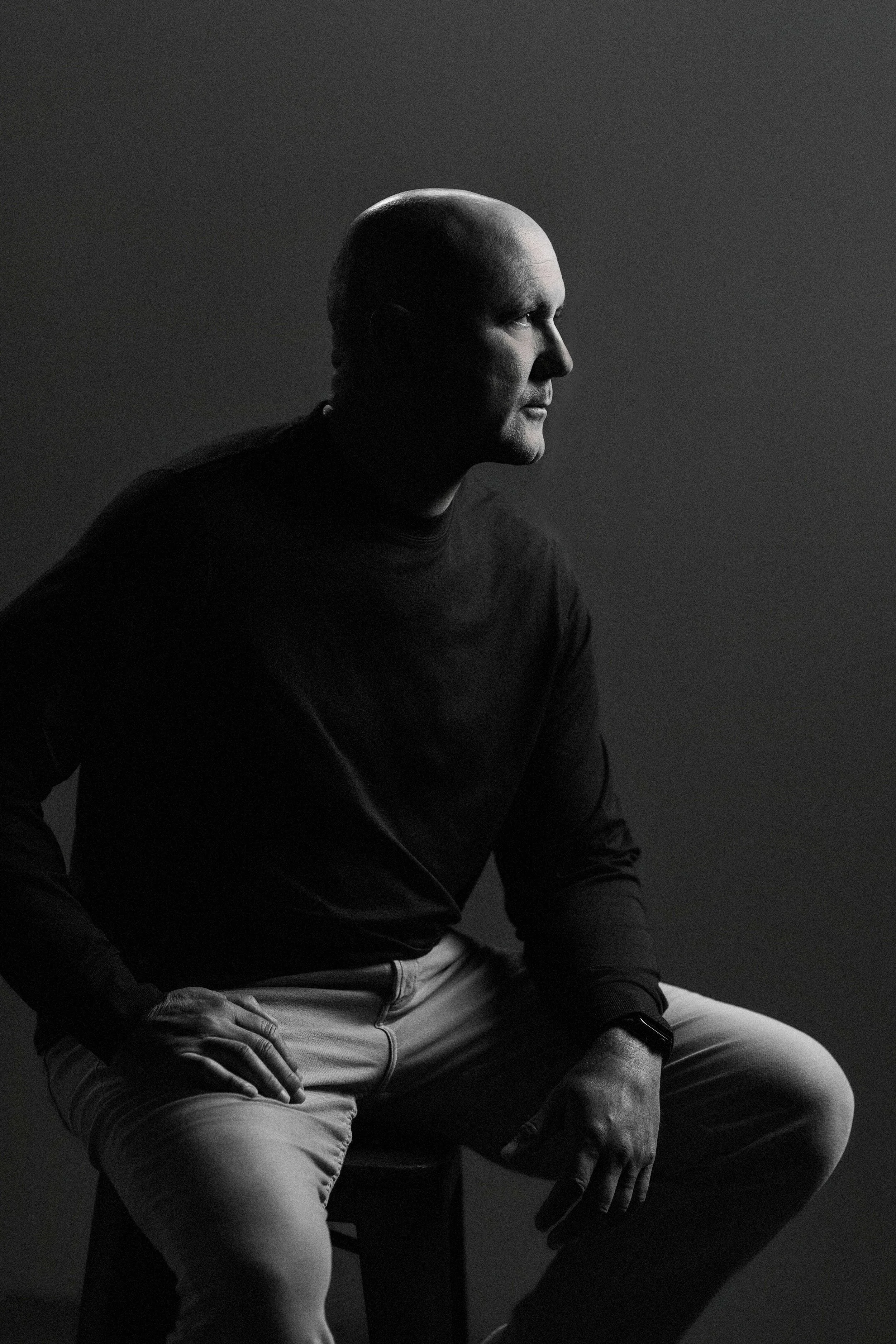 A black and white side portrait of a bald man sitting on a stool, wearing a dark shirt and light-colored pants, with one hand on his knee and the other resting on his thigh, against a plain dark background.