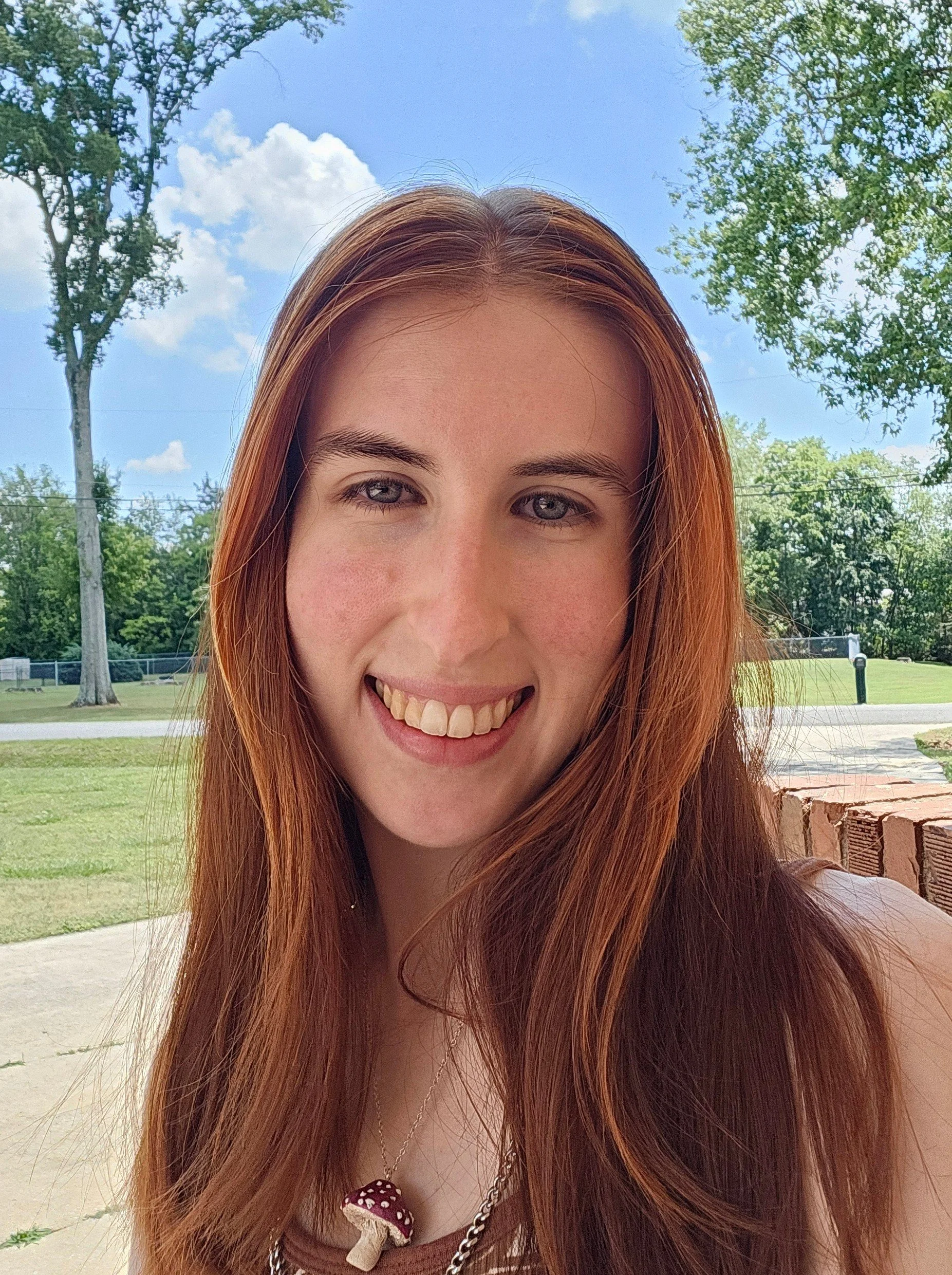 Ace of Huntsville Behavior Associates with long red hair, smiling outdoors with trees and blue sky in the background.