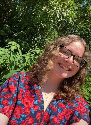 Amber of Huntsville Behavior Associates with curly hair and glasses, smiling in a floral dress outdoors among green foliage.