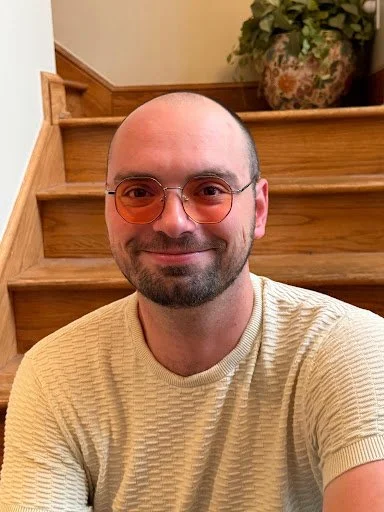 Jacob of Huntsville Behavior Associates wearing glasses and a beige sweater, smiling indoors near wooden stairs and a potted plant.
