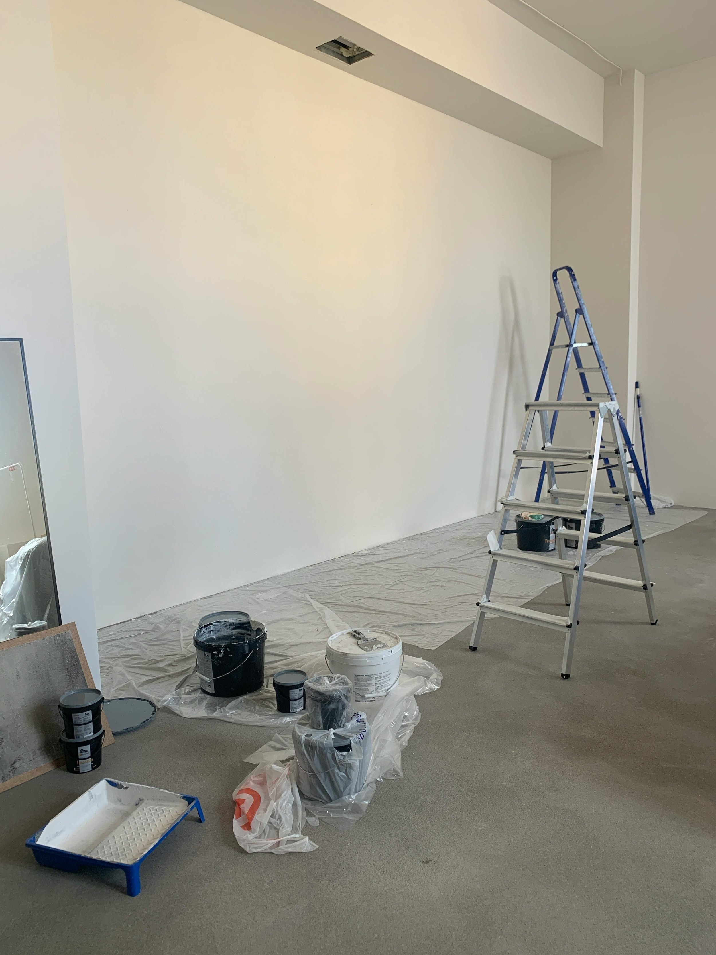 Inside a room under renovation with white painted walls, painting supplies scattered on the floor, including open paint cans, brushes, a paint roller, a small paint tray, and a step ladder. Part of the ceiling is drywalled with an opening for lighting or a vent.