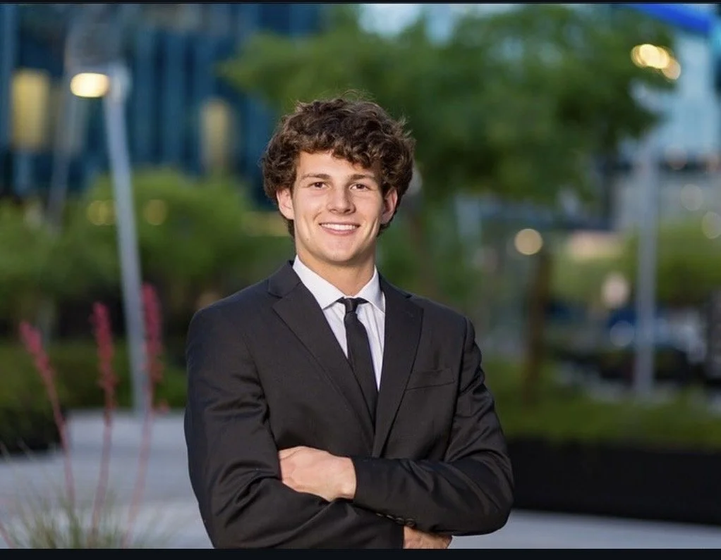 A young man with curly hair wearing a black suit, white shirt, and black tie, smiling with arms crossed outdoors in a park-like setting with greenery and blurred buildings in the background.