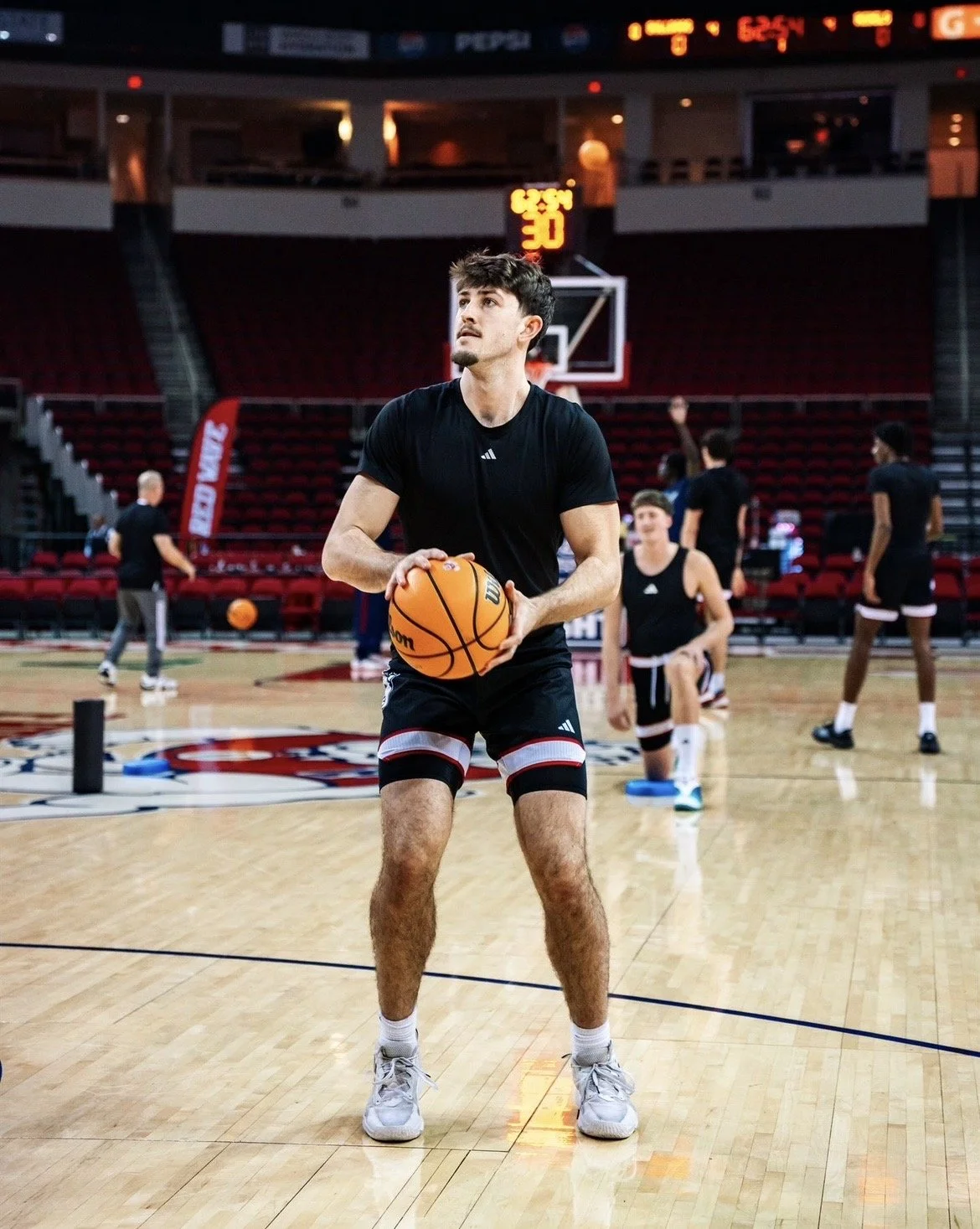 A young man in black Adidas sportswear preparing to shoot a basketball on an indoor court with several other young men practicing in the background.