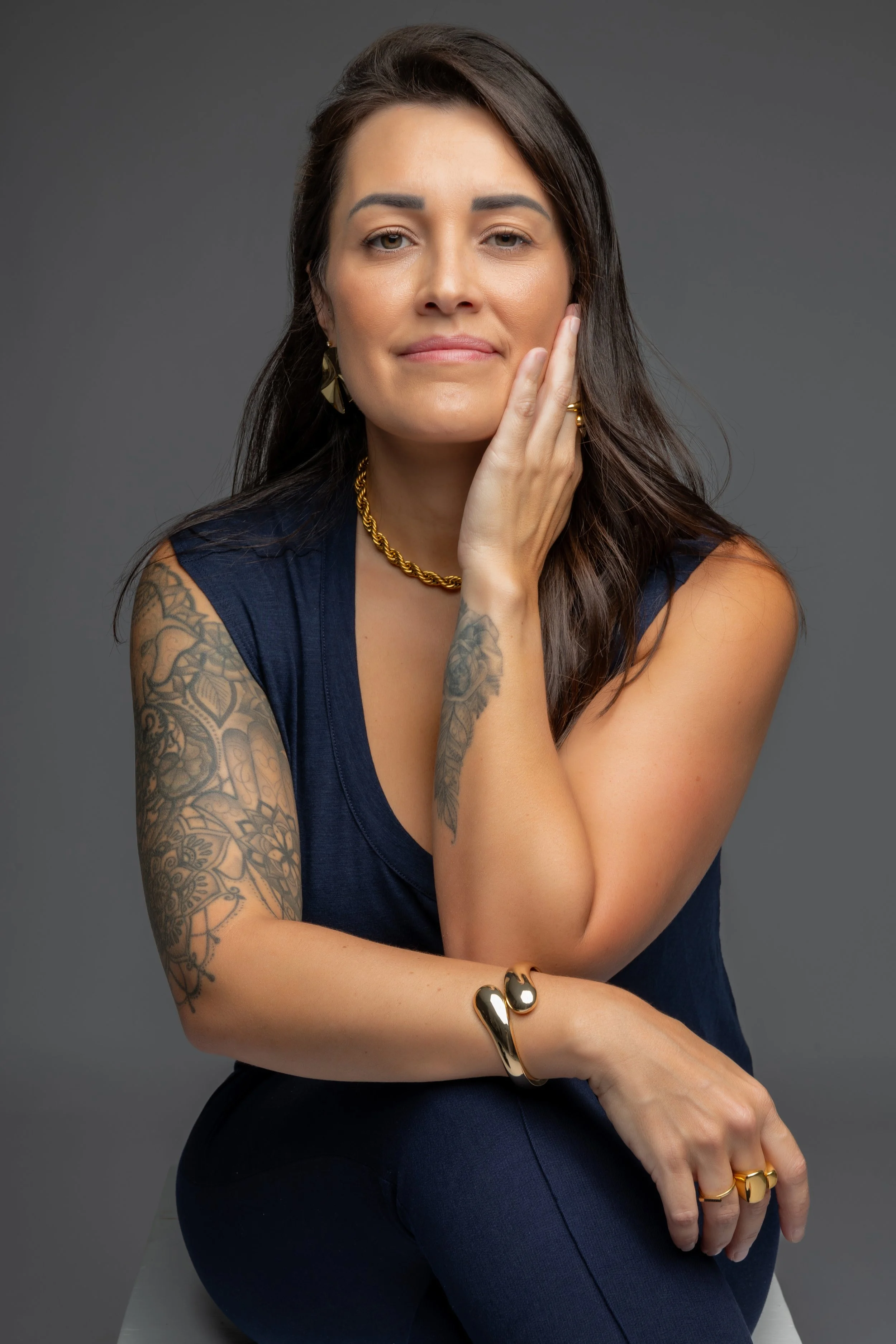 A woman with long dark hair, tattoos on her arms, wearing a navy blue sleeveless top, gold jewelry, and a gold bracelet, poses with her hand touching her face against a gray background.