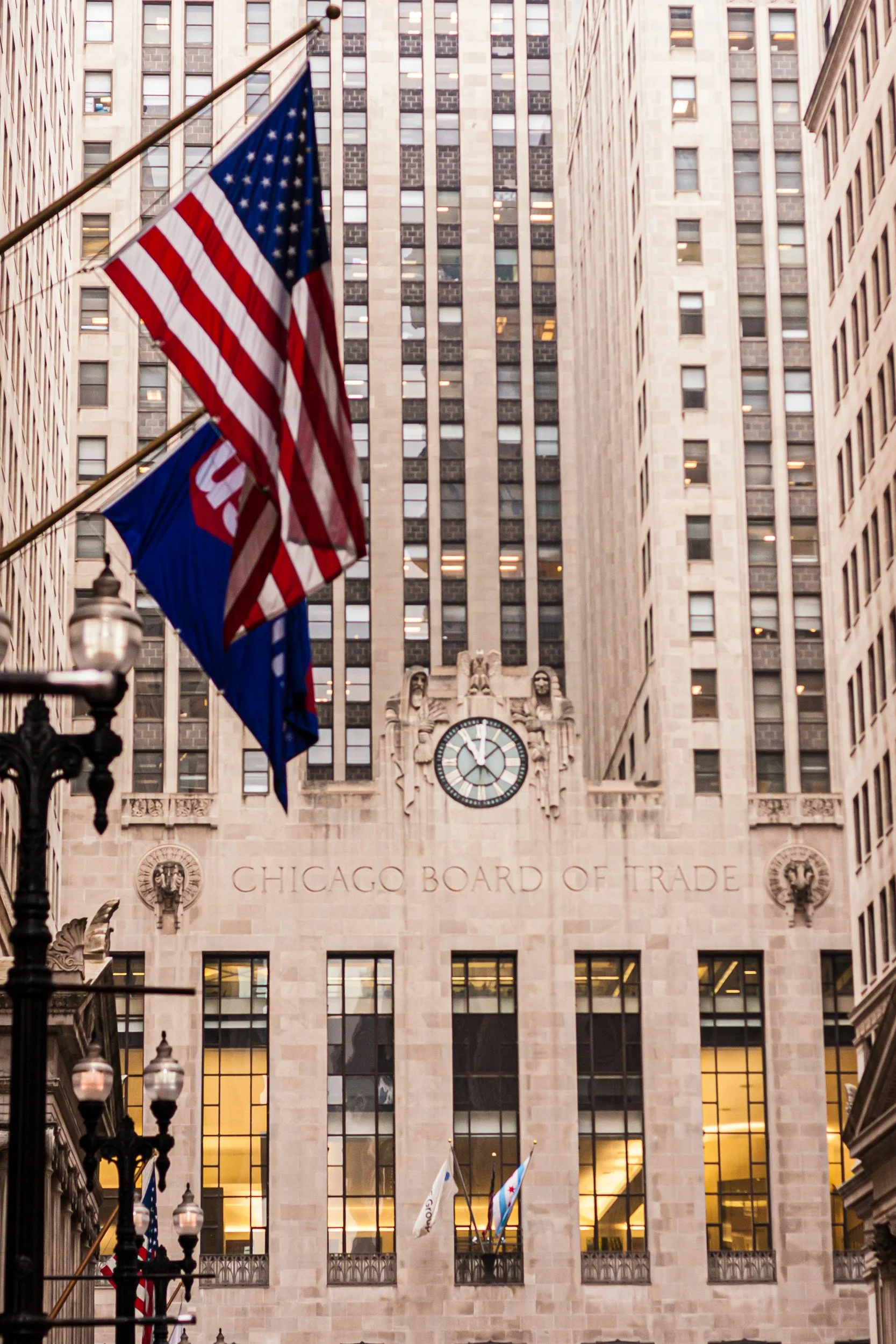 The front of the Chicago Board of Trade building with flags and a clock above the entrance, surrounded by tall office buildings.
