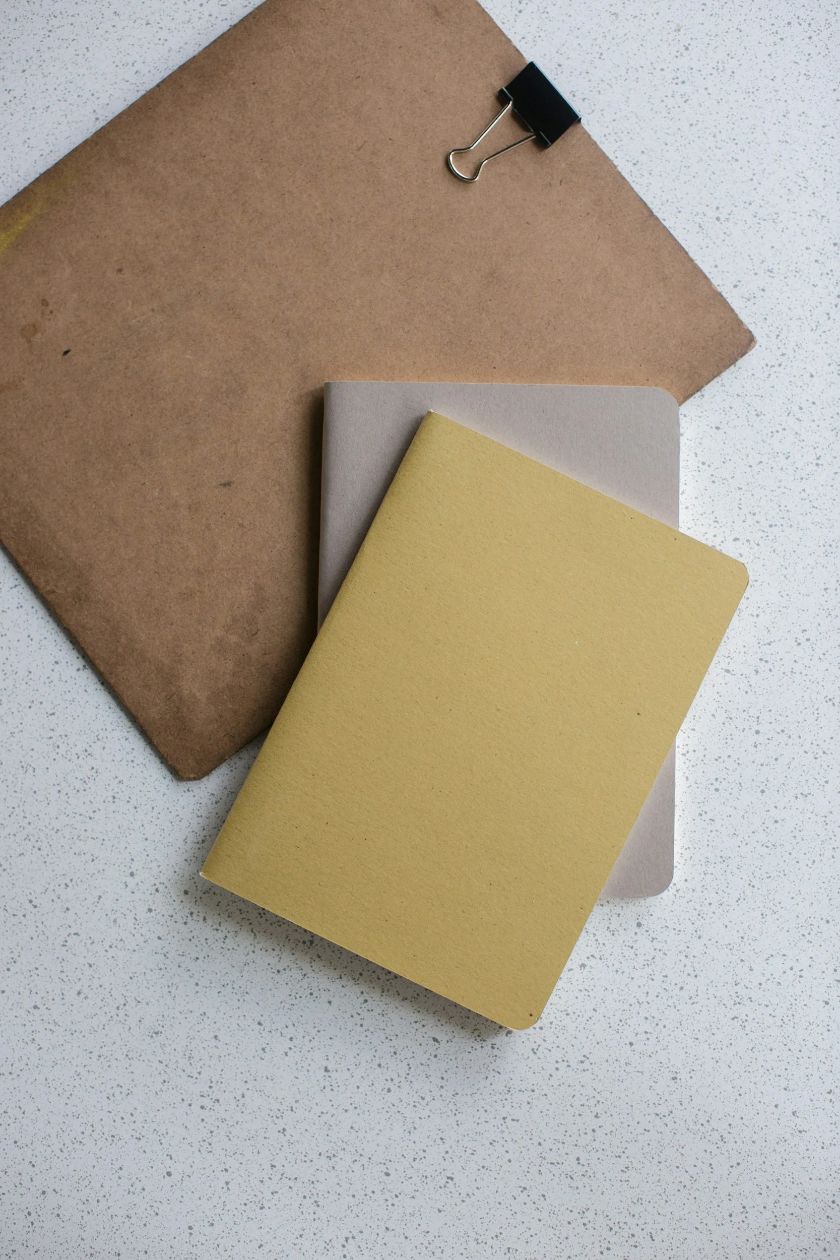 Flat lay of a brown cork notebook, a white notebook, and a yellow notebook on a speckled white surface, with a black binder clip attached to the cork notebook.