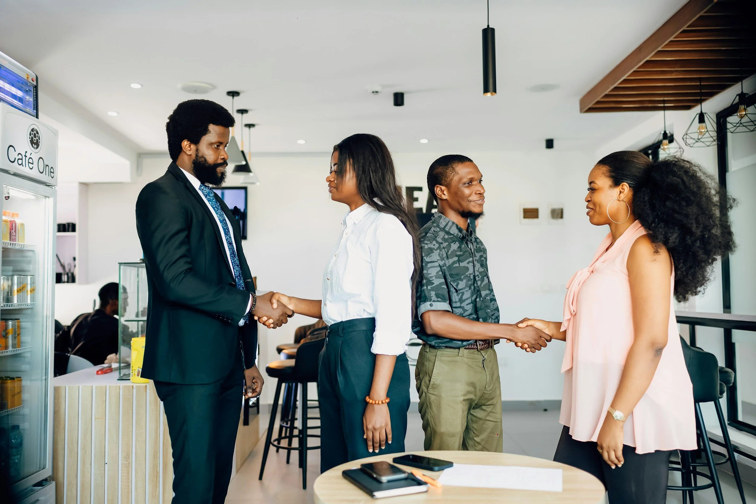 A group of four diverse people shaking hands in a modern, bright cafe or office space. Two men and two women are standing in a line, exchanging handshakes with smiles. There are tables, chairs, and a drink fridge in the background.
